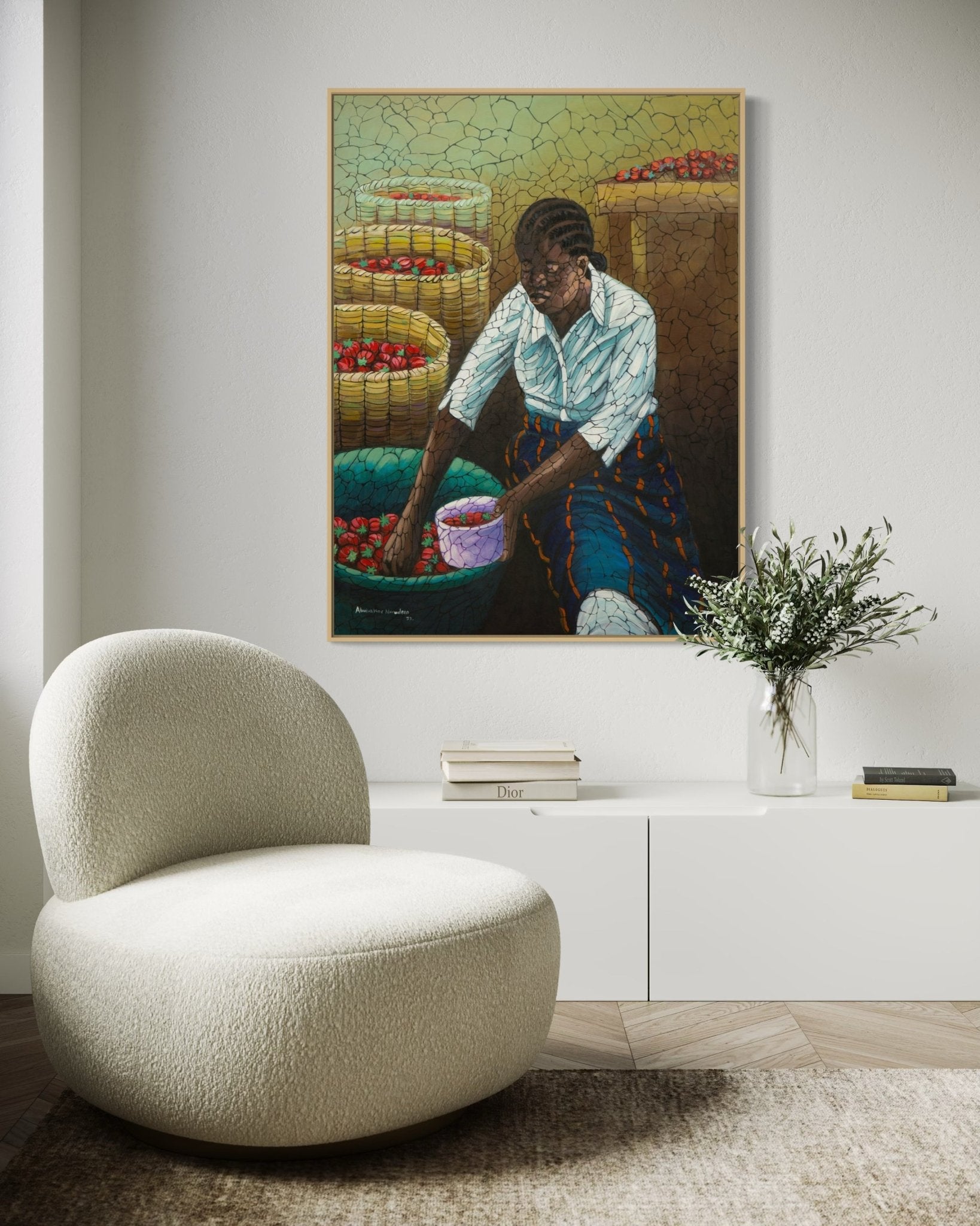 A modern living room with a curved cream chair, a low white cabinet, a vase with greenery, and a framed painting of a woman sorting tomatoes in baskets hanging on the wall above.