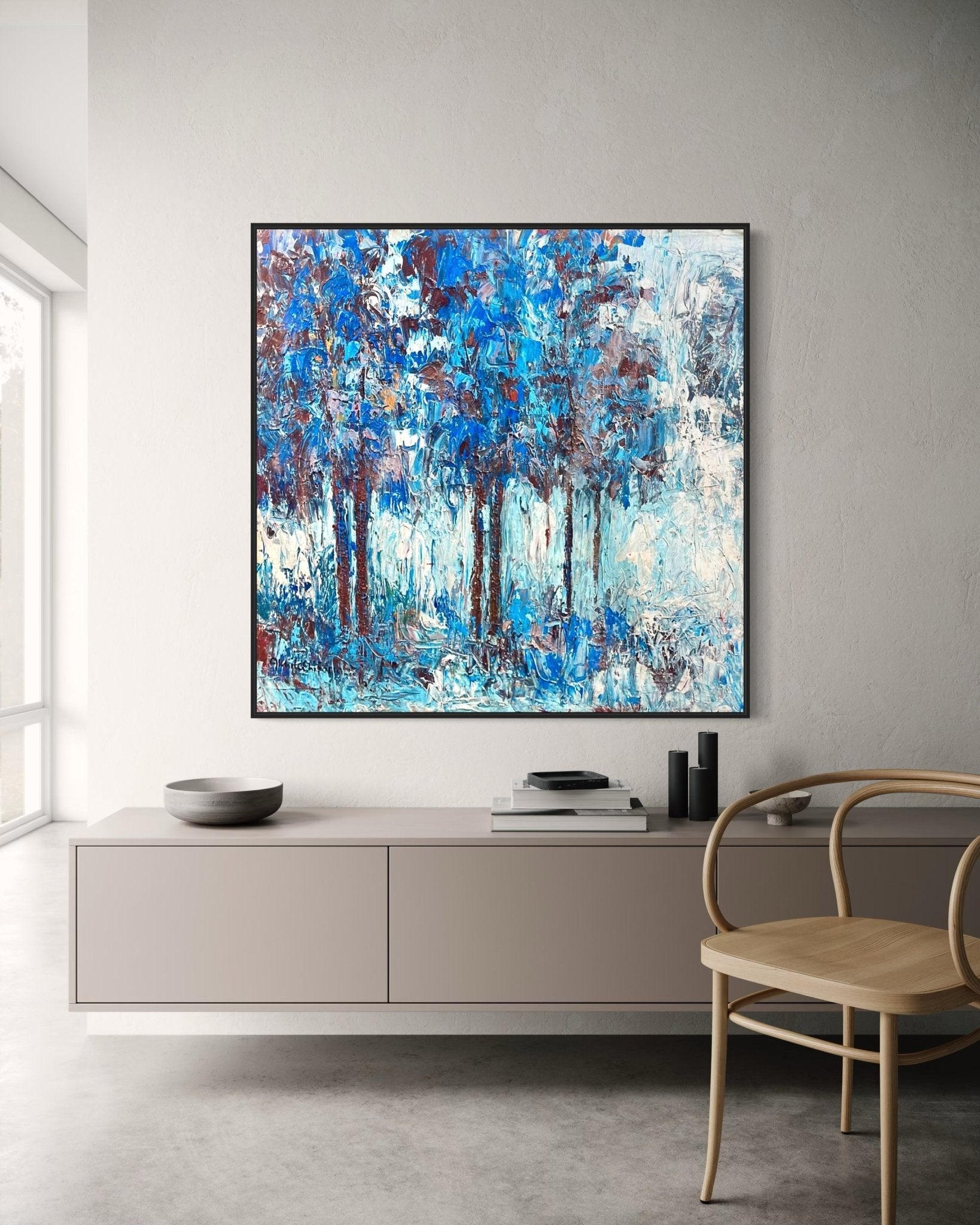 A minimalist room with light walls, a wooden chair, a low cabinet, and a large abstract blue and white painting of trees hanging above the cabinet. Natural light enters from a window on the left.