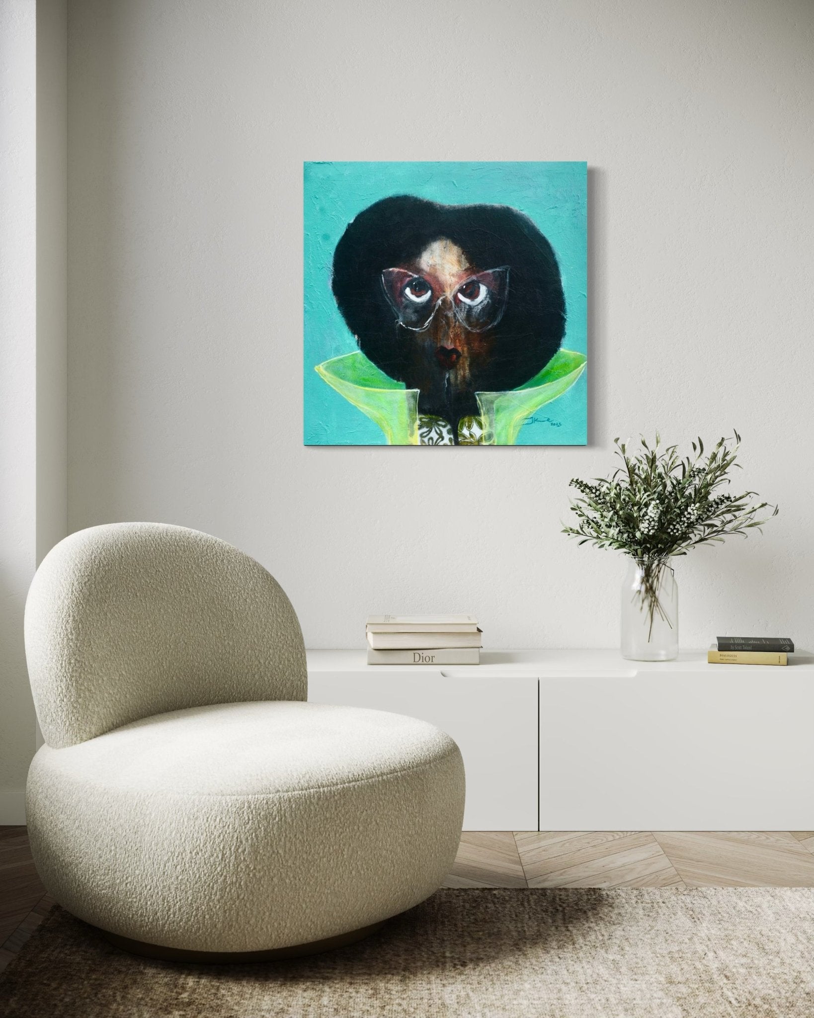 A modern living room with a curved beige chair, a low white sideboard with books, a clear vase with greenery, and a colorful painting of an abstract face on the white wall above.