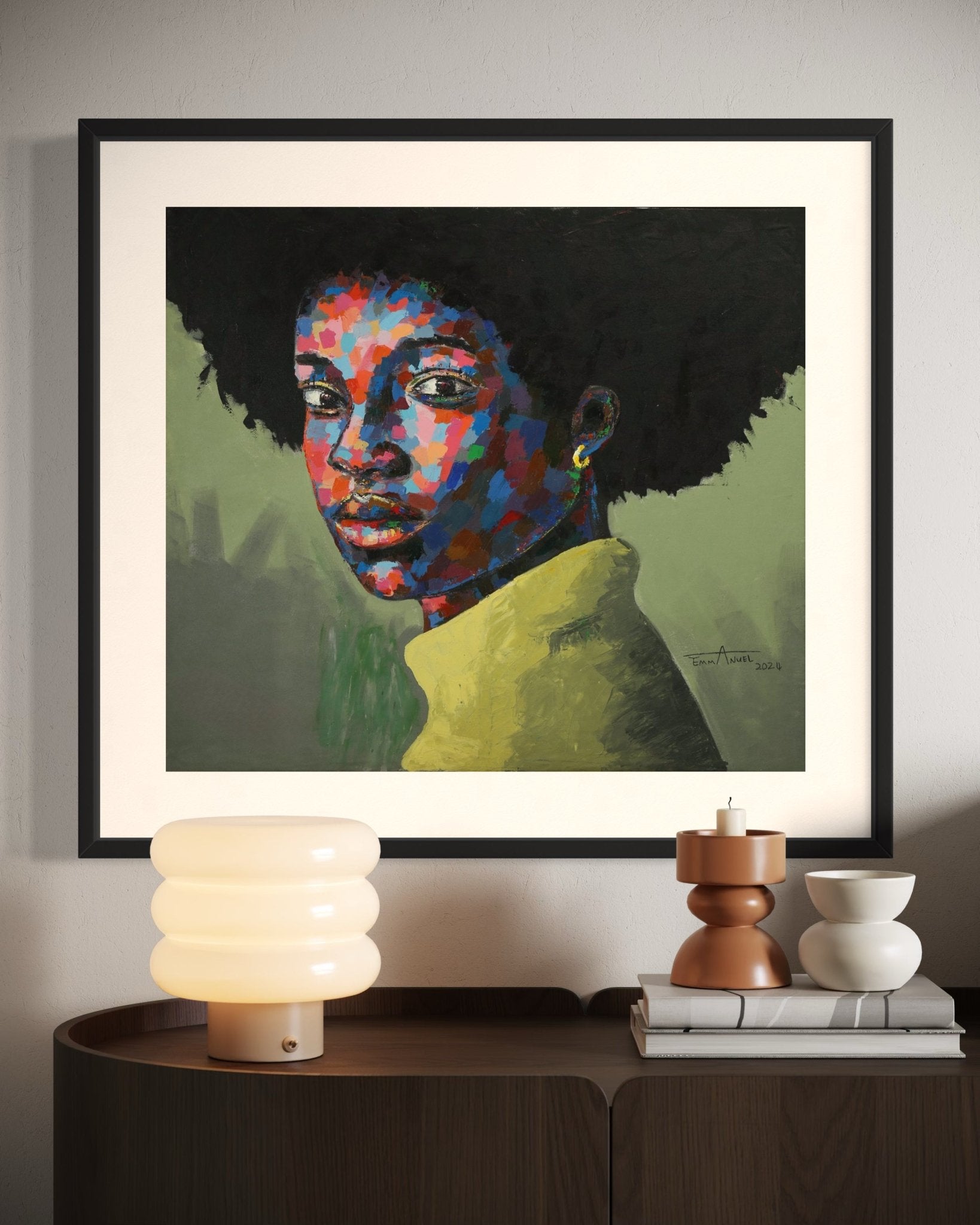 A framed abstract portrait of a woman with an afro hangs above a modern console table with a lamp, stacked books, a candle, and a bowl. The painting features bold, colorful brushstrokes.
