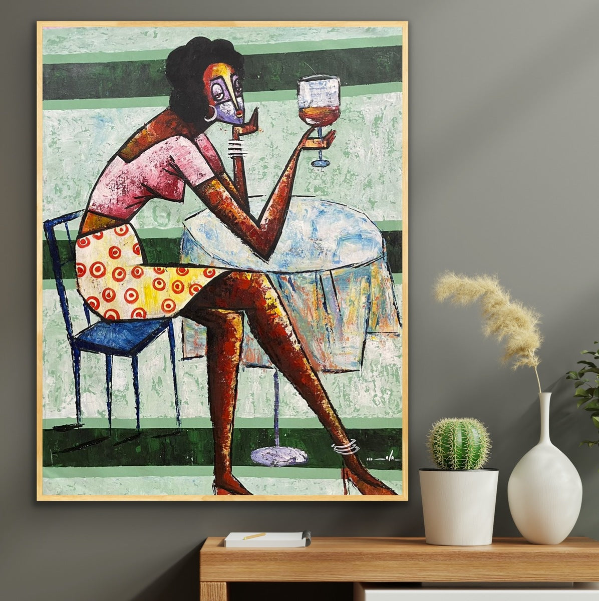 Cheers" by Ola Kuti is a vibrant acrylic on canvas of a dark-skinned woman with curly hair, seated on a blue chair at a table with wine. Displayed above a shelf with plants, this artwork is hand-signed by the artist.