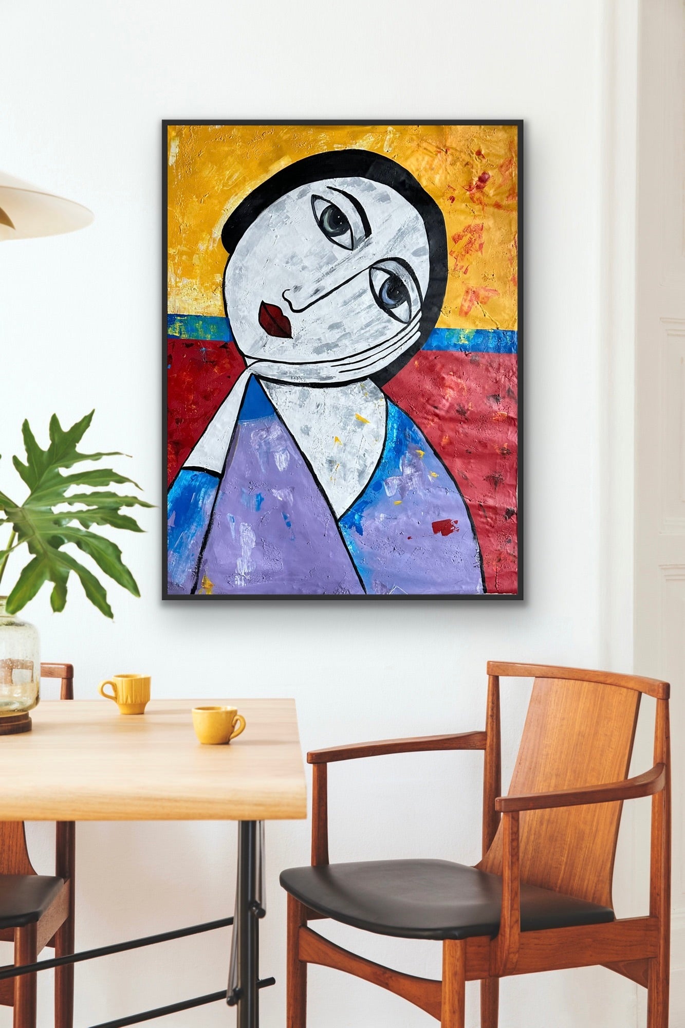 The Ifeanyi Admire framed abstract painting by Ifeanyi Admire hangs on a white wall above a wooden dining table with two yellow cups, a green plant, and wooden chairs.
