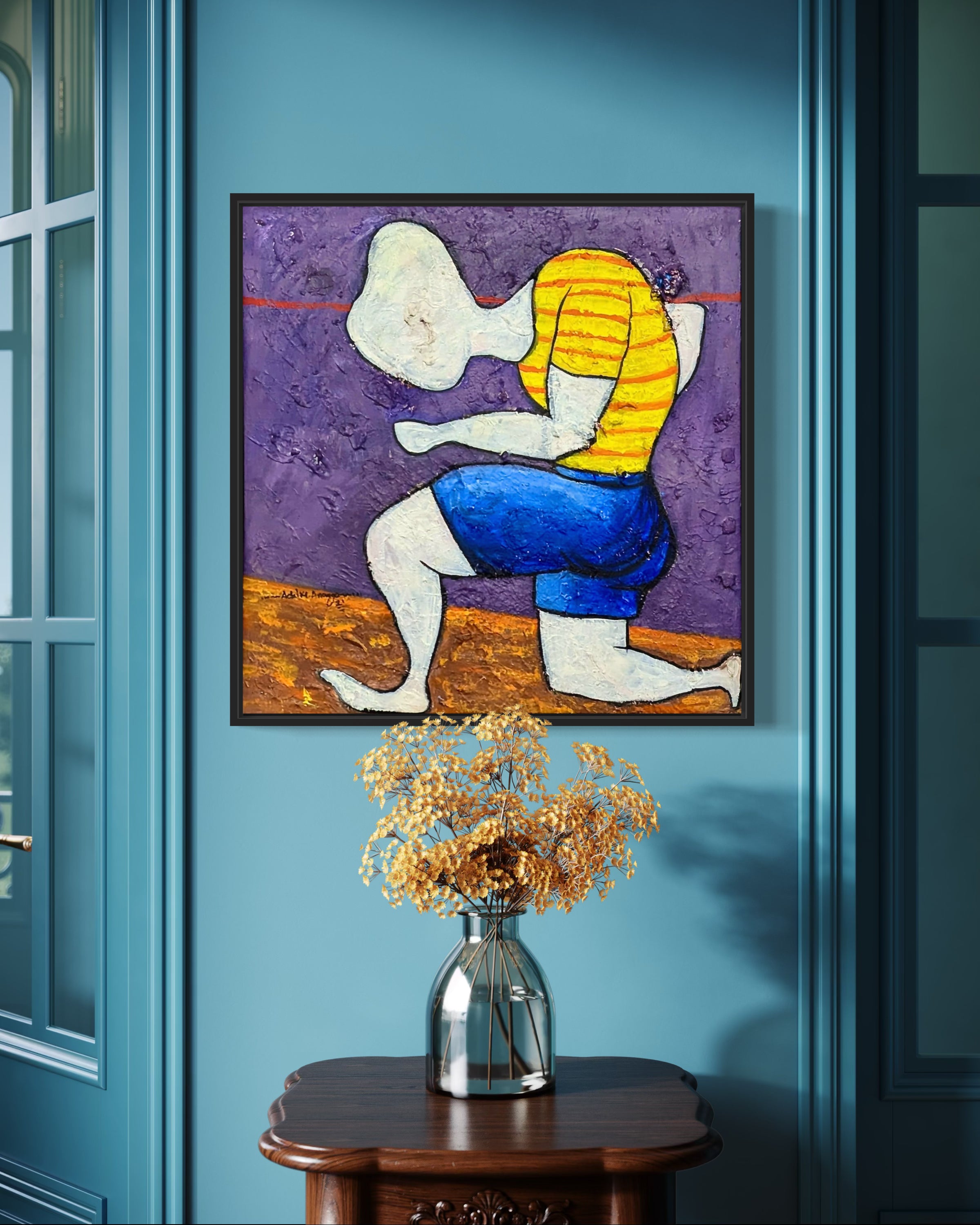 Dance Of Nature" by Achike Anayo, an expressionist painting of an abstract figure in a yellow striped shirt and blue shorts, is hand-signed and displayed in a black tray frame on a blue wall above a wooden table with dried flowers.