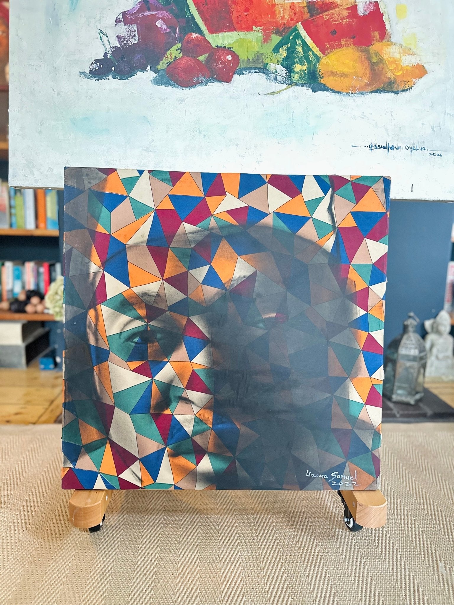 Uzoma Samuel's Abstract Figurative portrait "Self" features vibrant geometric triangles forming a stylized face, displayed upright on a wooden stand with bookshelves and artwork in the background.
