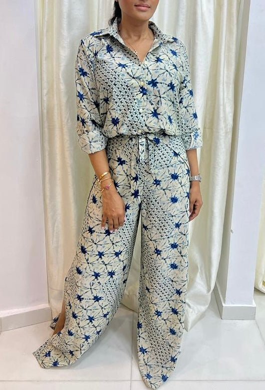 A woman stands indoors wearing the Adire Batik Chiffon Multiway 2 Piece Top & Wrap Pant Set by bibianna.co.uk, featuring a blue and black starburst pattern on white. Her sleeves are rolled up and she accessorizes with bracelets.