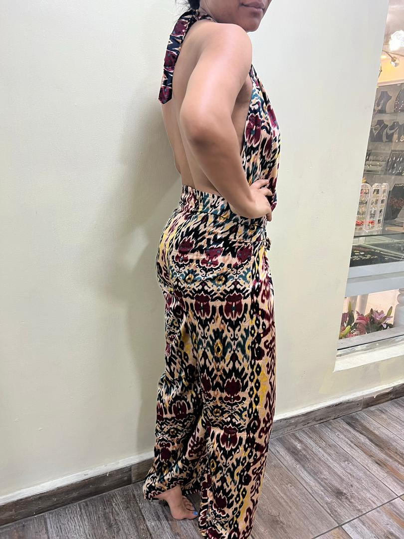 A person stands barefoot indoors near a wall and window, wearing the vibrant Adire Batik Chiffon Multiway 2 Piece Top & Wrap Pant Set from bibianna.co.uk—a colorful, patterned co-ord from an African label.