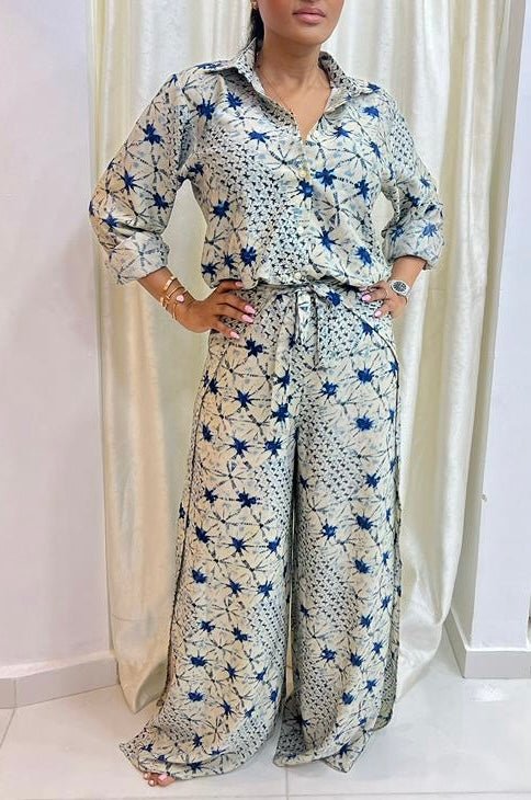 A woman stands with hands on hips in the Adire Batik Chiffon Multiway 2 Piece Top & Wrap Pant Set from bibianna.co.uk, a cream co-ord with blue star patterns, collared button-up shirt, wide-leg pants, and gold accessories.