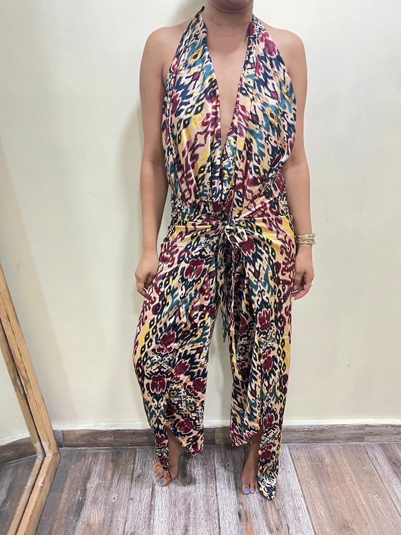 A person stands indoors wearing the Adire Batik Chiffon Multiway 2 Piece Top & Wrap Pant Set by bibianna.co.uk, featuring colorful abstract red, yellow, blue, and black designs. The face is not visible.