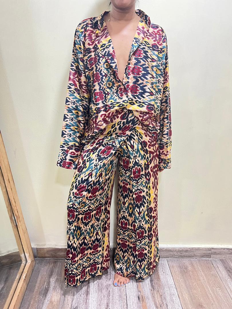 Indoors, a person wears the Adire Batik Chiffon Multiway 2 Piece Top & Wrap Pant Set from bibianna.co.uk, featuring bold red, yellow, blue, and black geometric and abstract prints by an African label.
