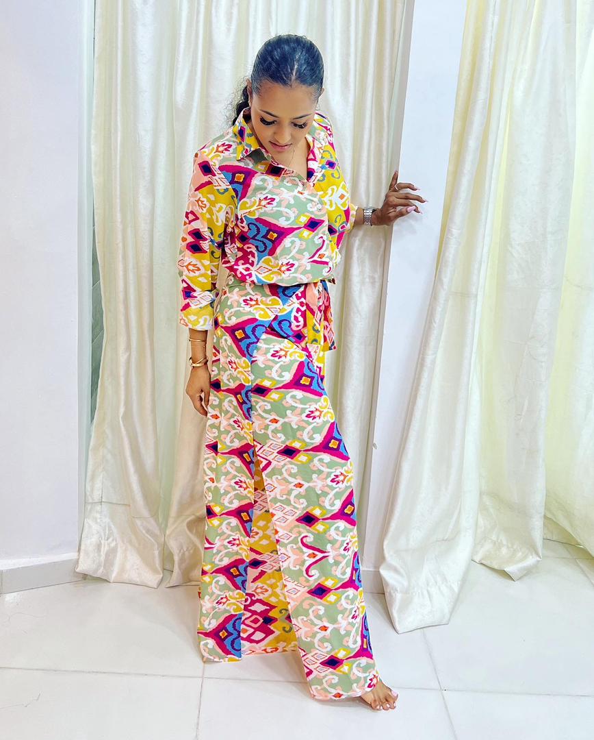 A woman stands barefoot indoors, wearing the Adire Batik Chiffon Multiway 2 Piece Top & Wrap Pant Set from bibianna.co.uk. She looks down gently, holding a curtain and posing in front of white drapes on a tiled floor.