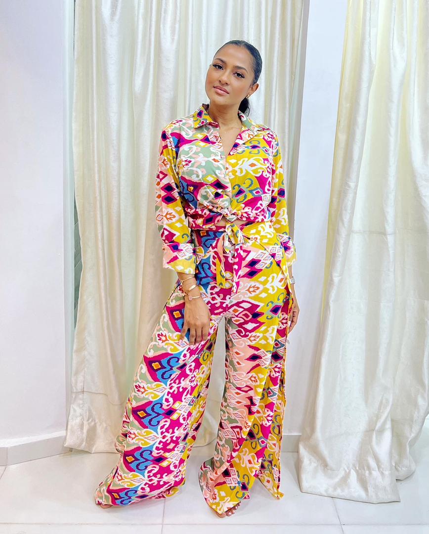 A woman stands indoors in bibianna.co.uk's Adire Batik Chiffon Multiway 2 Piece Top & Wrap Pant Set, featuring colorful pink, yellow, green, and white patterns. She poses confidently before white curtains in the vibrant African-inspired outfit.
