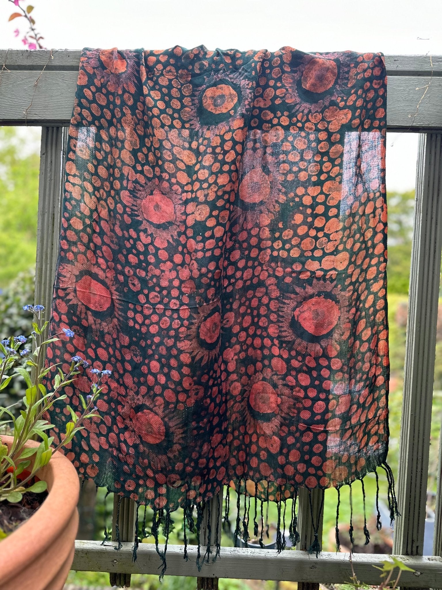 The Adire Pashmina Scarf - Mina by bibianna.co.uk, featuring unique circular red and orange patterns, hangs over a wooden balcony railing. A potted plant with blue flowers sits nearby, with greenery in the background.