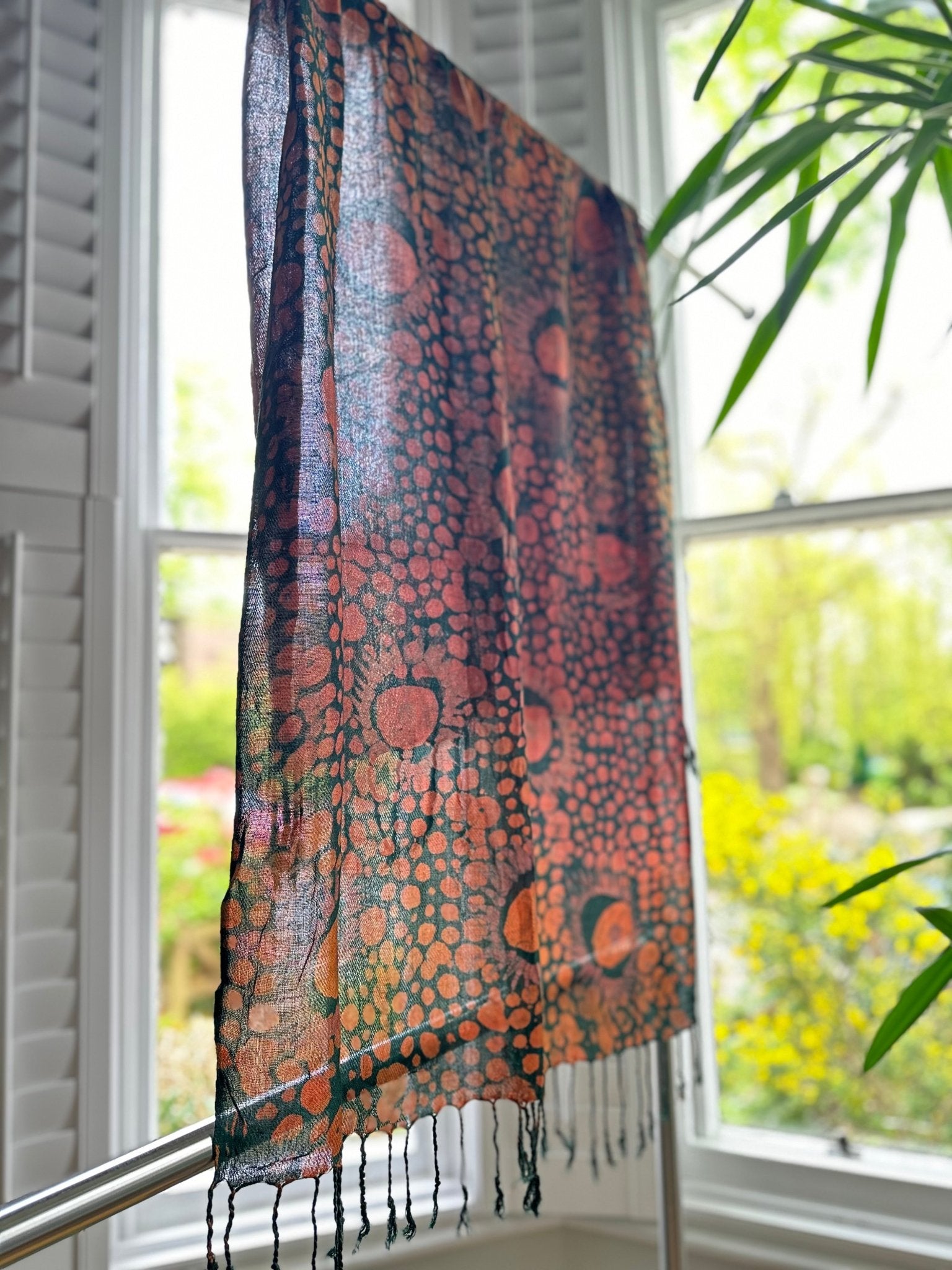 The Adire Pashmina Scarf - Mina by bibianna.co.uk, a sheer handmade scarf with a unique circles and dots pattern, hangs on a rack in front of large windows with white shutters, greenery and trees visible outside.