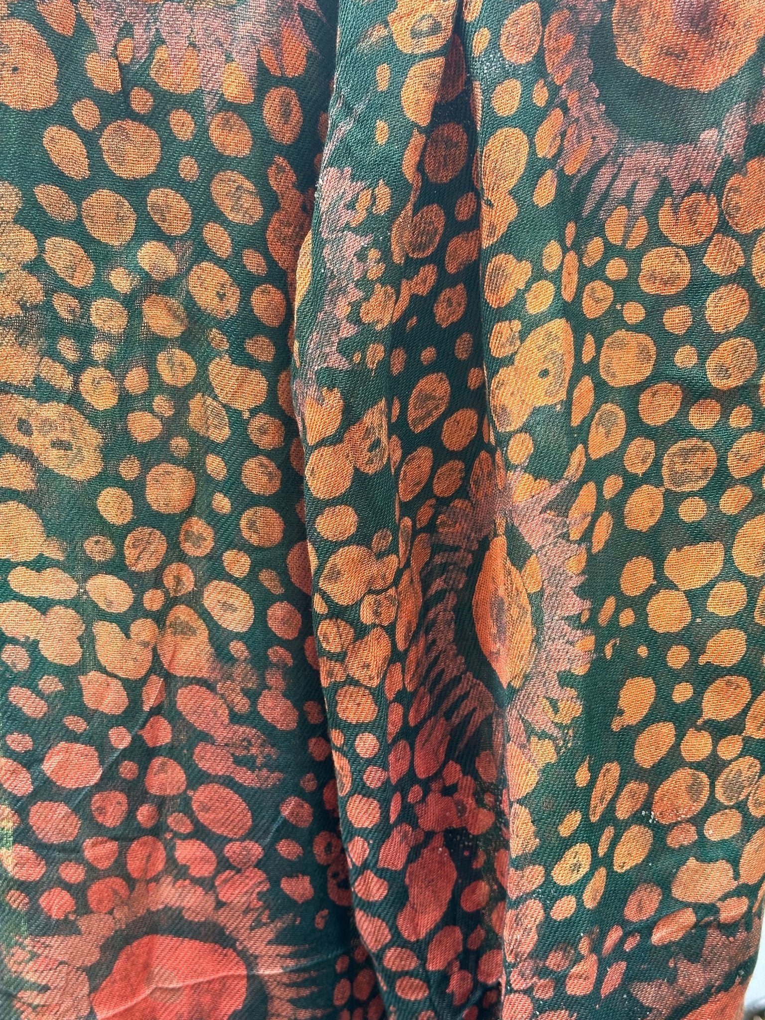 The Adire Pashmina Scarf - Mina by bibianna.co.uk features a handmade sunburst pattern of orange and yellow dots and circles on dark green, with visible folds and texture for added charm.