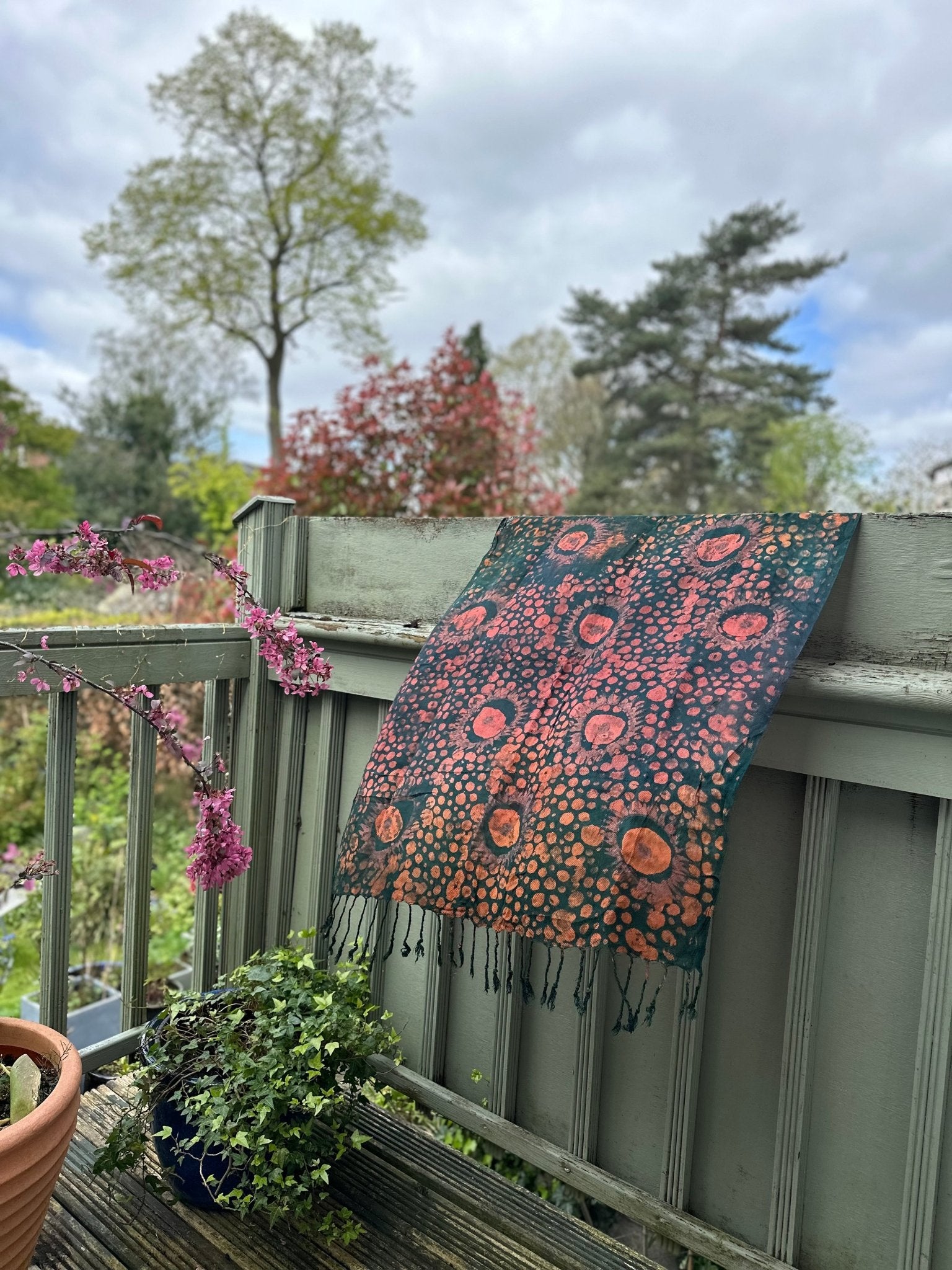 The Adire Pashmina Scarf - Mina by bibianna.co.uk, featuring unique orange and pink circular and dotted patterns, hangs over a pale green porch railing with potted plants and blooming trees under a cloudy sky.