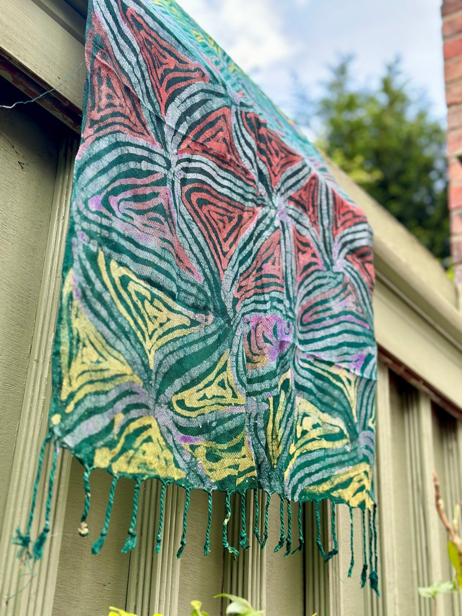 The Adire Pashmina Scarf - Star by bibianna.co.uk features a vibrant handmade geometric pattern in red, yellow, green, and blue with tassels, displayed draped over a wooden fence against a backdrop of blue sky and greenery.