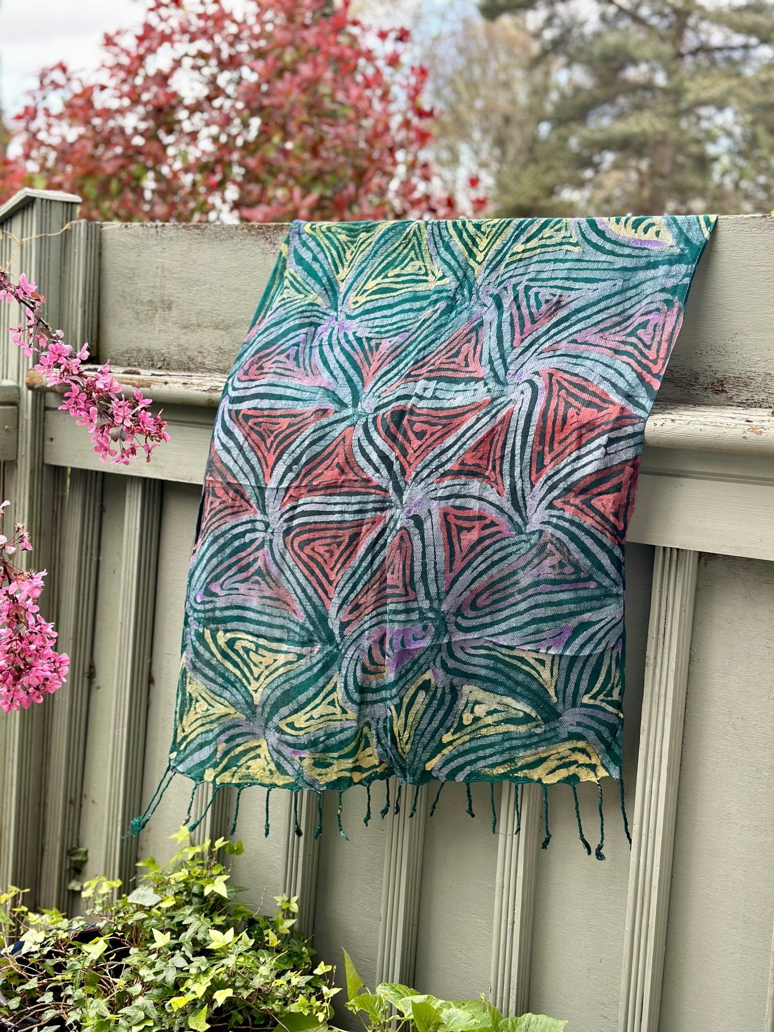 The Adire Pashmina scarf - Star by bibianna.co.uk, featuring a handmade geometric leaf pattern in green, pink, blue, and yellow, hangs over a light green fence beside blooming pink flowers and lush green foliage.