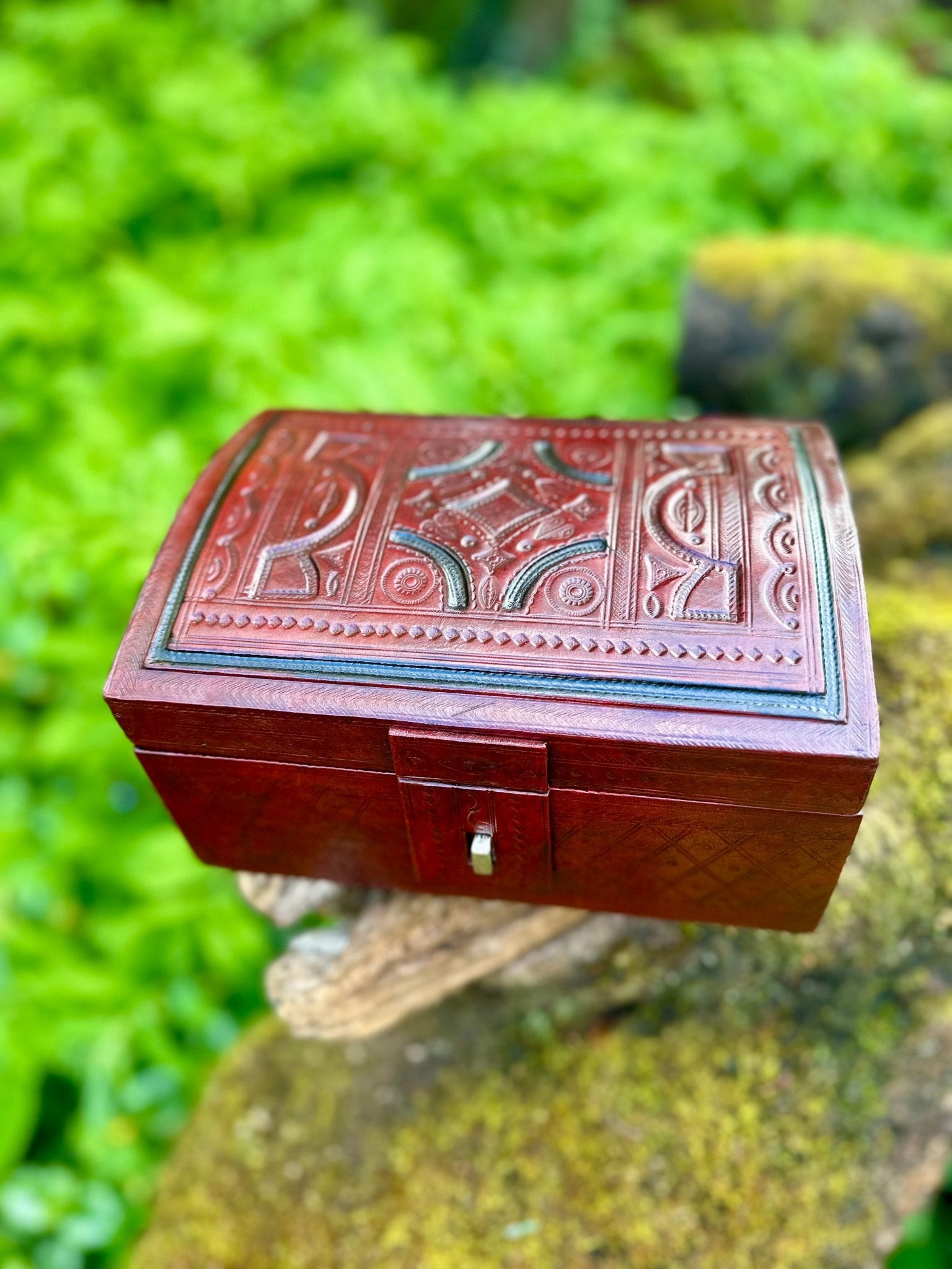 The African Tuareg Style Hand-Tooled Red Leather Box by bibianna.co.uk rests on a mossy rock outdoors, its intricate detailing highlighted against a backdrop of lush green foliage.