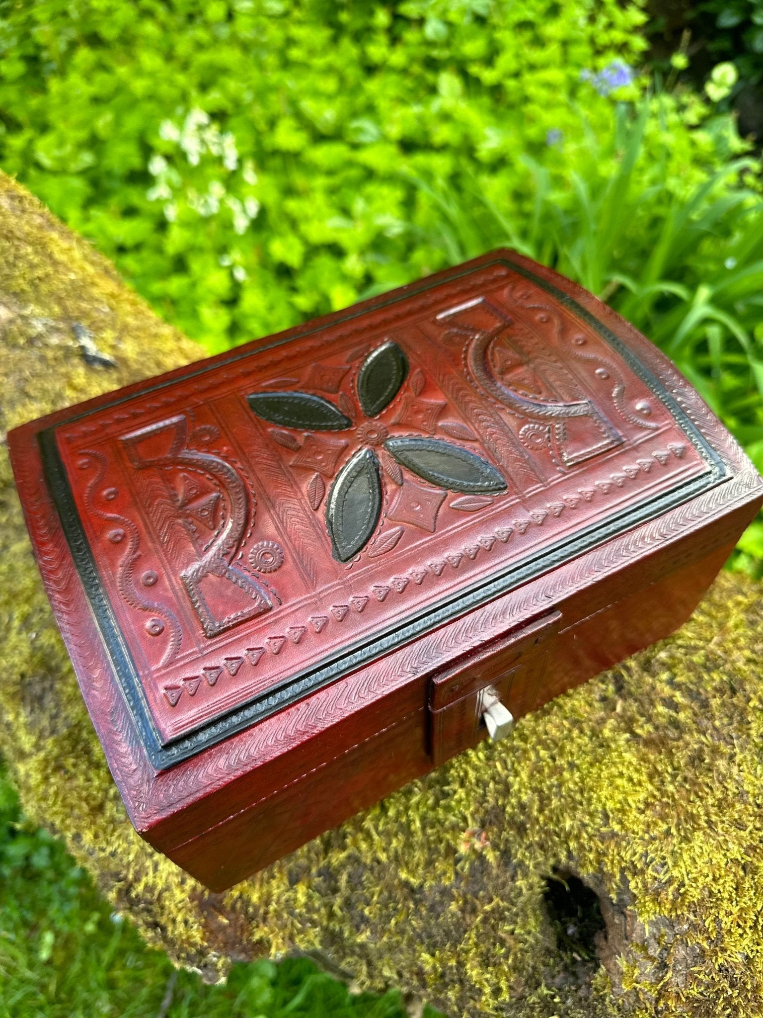 The African Tuareg Style Hand-Tooled Red Leather Box by bibianna.co.uk, featuring intricate patterns, rests outdoors amid vibrant greenery—an ideal choice for distinctive ethnic home decor.