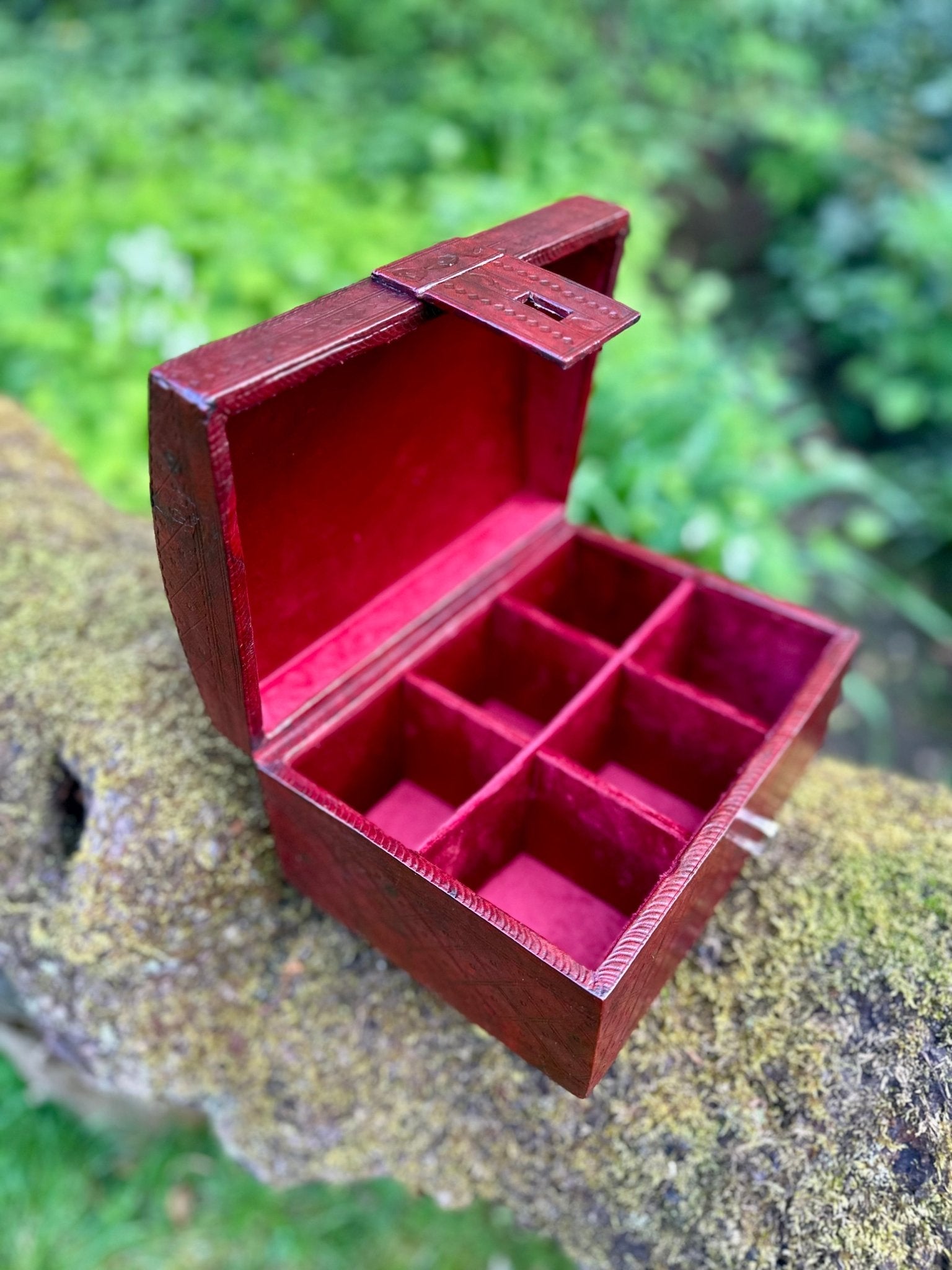 The African Tuareg Style Hand-Tooled Red Leather Box by bibianna.co.uk, featuring six square compartments lined in red velvet, is displayed outdoors on a mossy tree branch amidst lush green foliage.