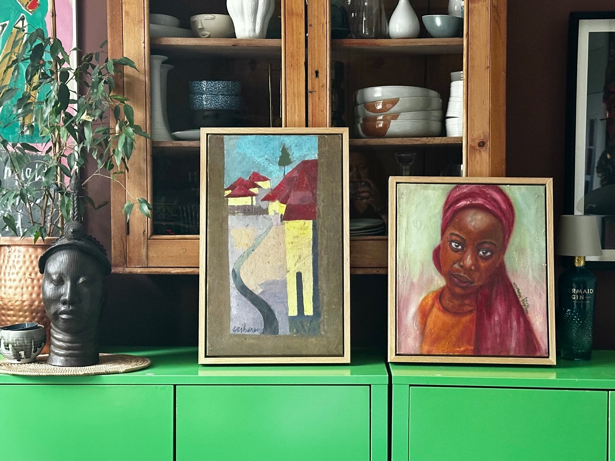 Two framed artworks—a hand-signed abstract landscape and "Little Amina," a Bibianna African Art Collective portrait of a woman in an orange top—rest on a bright green cabinet, with shelves of dishes, a sculpted head, and a potted plant behind.