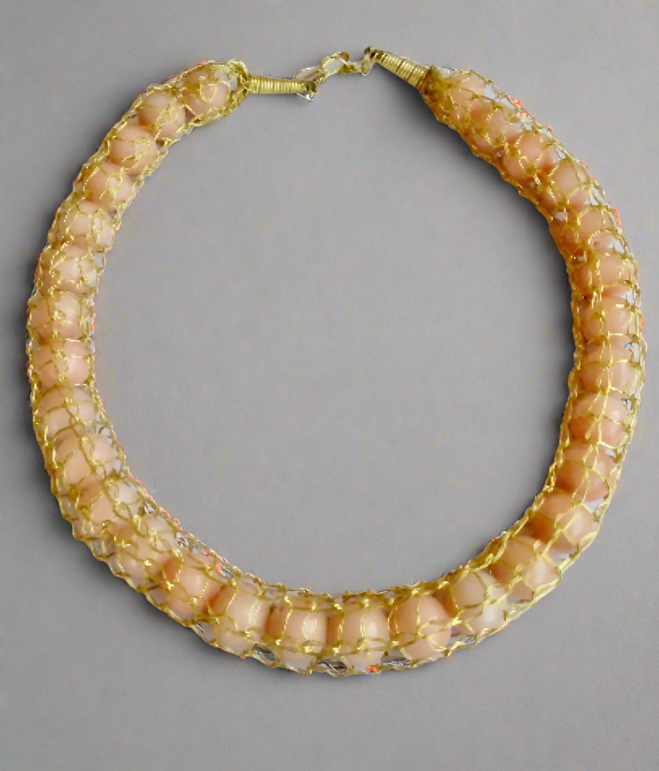 The Ariaba Crochet Coral Necklace by bibianna.co.uk features large oval pale pink coral stones in 14k gold-plated mesh, crafted in a circular design. Created by a West African designer, shown against a light gray background.