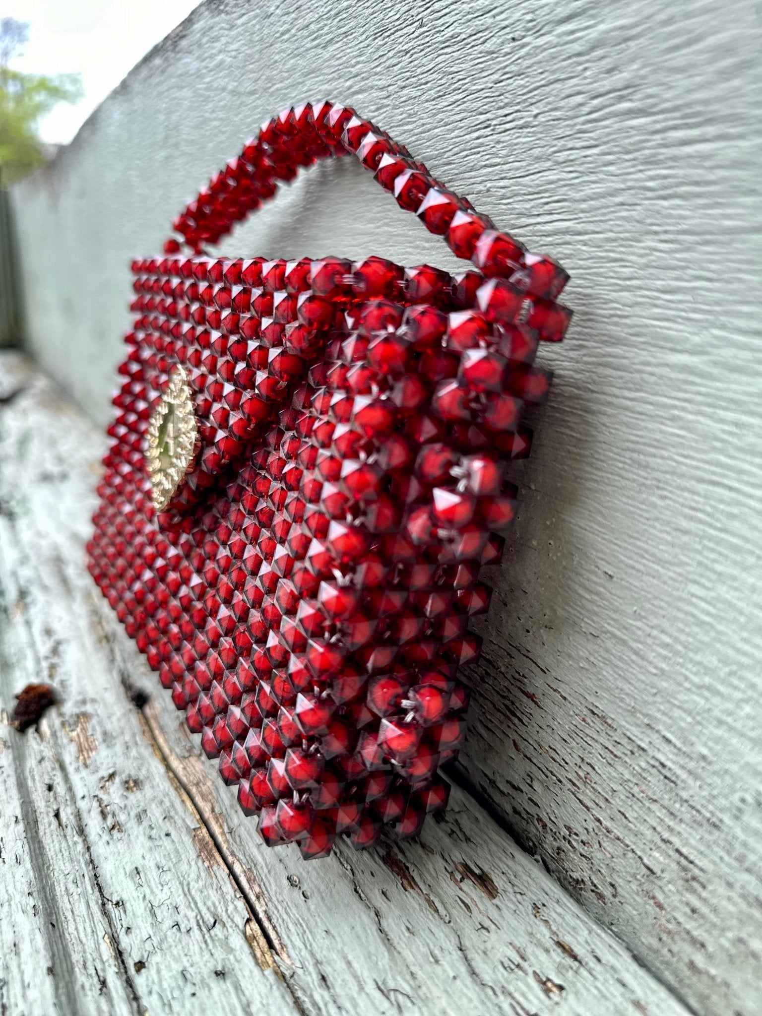 The Ariaba Hand Beaded Clutch by bibianna.co.uk, featuring intricate red beadwork and a gold clasp, sits on weathered wood against a pale gray wall, photographed from a low angle.