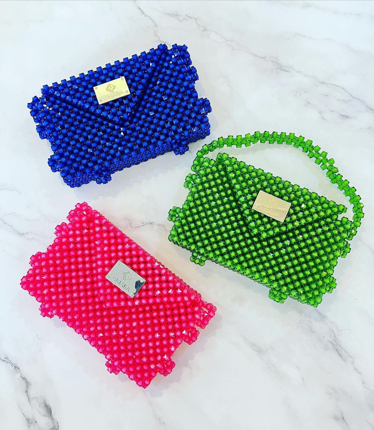 Three Ariaba Hand Beaded Clutch bags by bibianna.co.uk—blue, green (with beaded handle), and pink—showcase intricate beadwork, metallic labels, and are handmade in Nigeria. Displayed on a white marble surface.