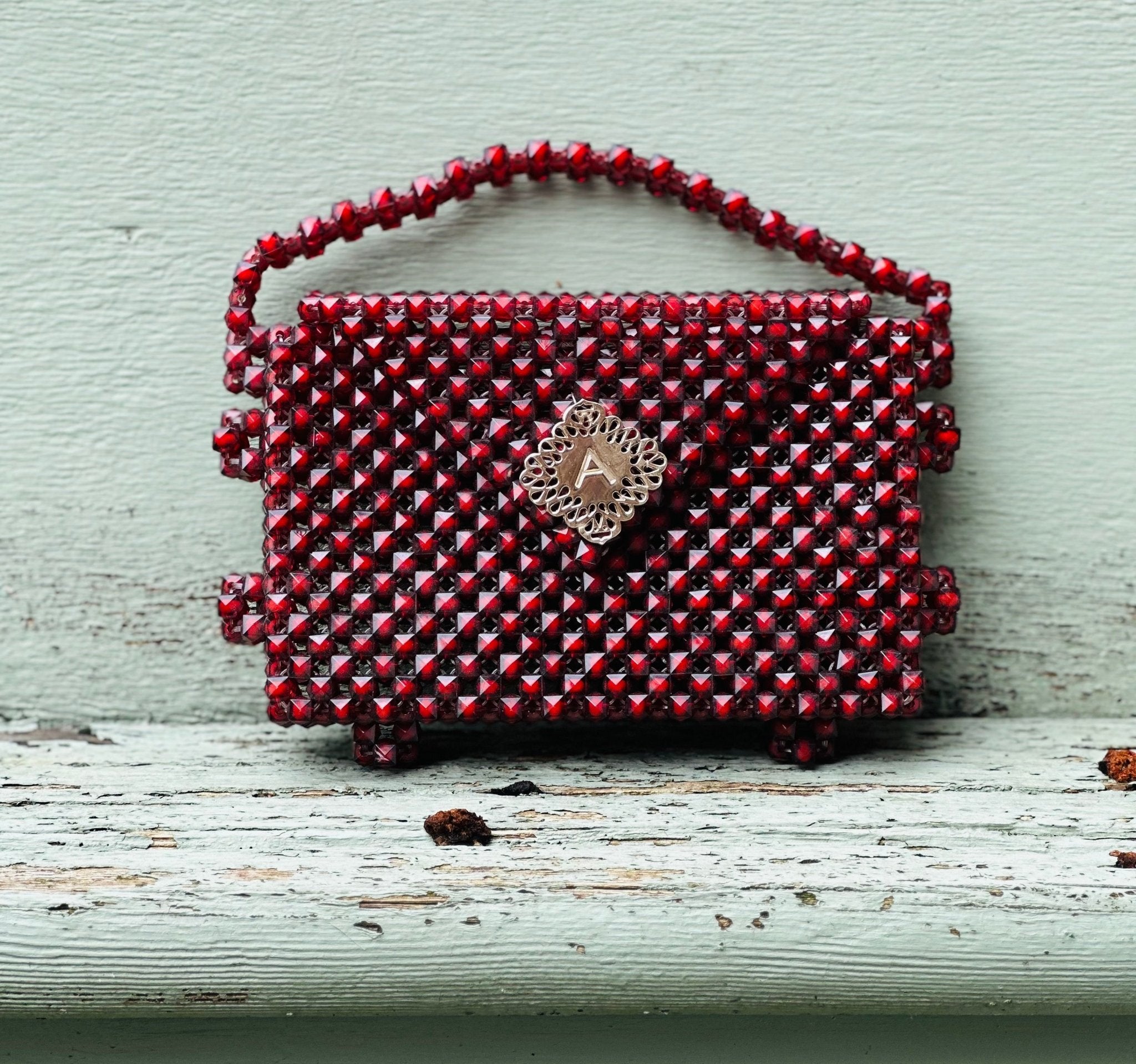 The Ariaba Hand Beaded Clutch from bibianna.co.uk is a small, rectangular red bag with a handle, shiny beadwork, and a silver clasp at the center. Handmade in Nigeria, it rests on a weathered white wooden surface.