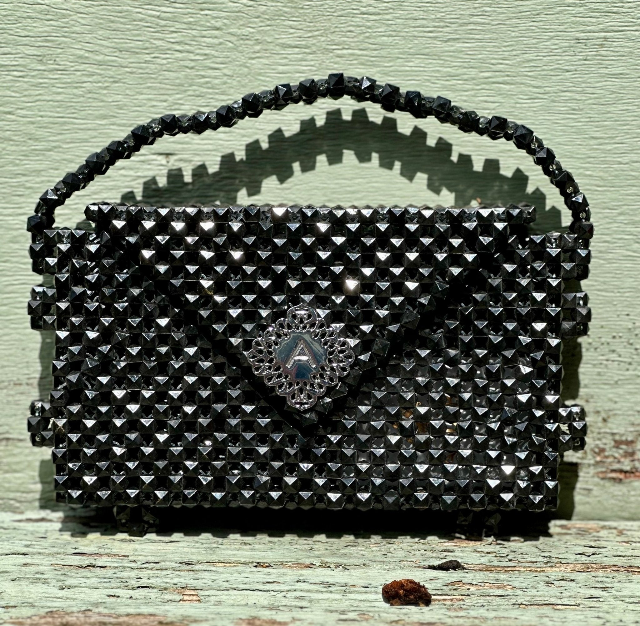 The Ariaba Hand Beaded Clutch by bibianna.co.uk, a small black purse with a triangular flap, shiny studs, and a silver clasp, sits on weathered green wood. This exquisite handmade piece showcases Nigerian beadwork.