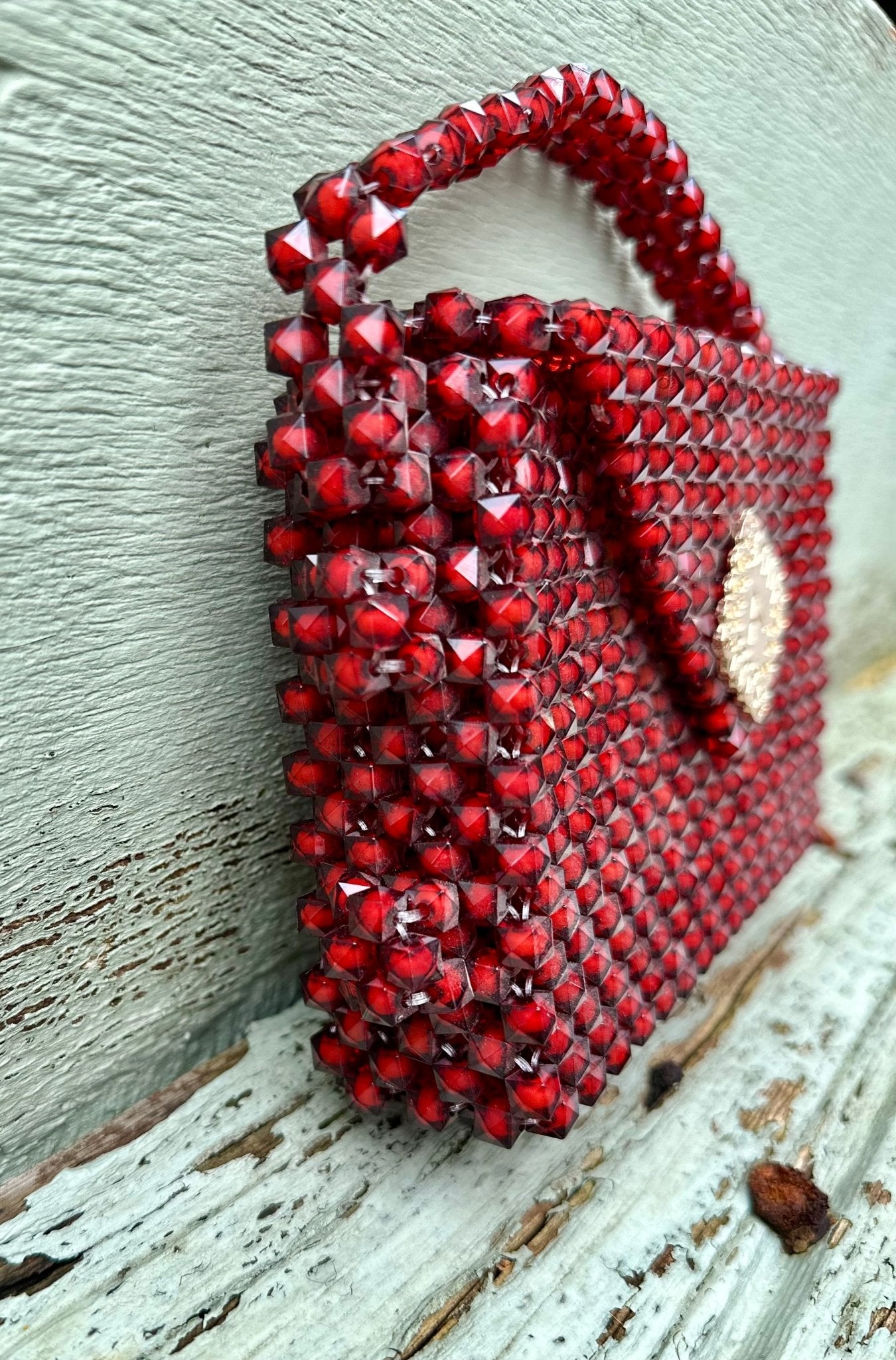 The Ariaba Hand Beaded Clutch by bibianna.co.uk is a small red clutch with intricate hand-beaded diamond accents and woven texture, showcased on a weathered wooden surface. Handmade in Nigeria.