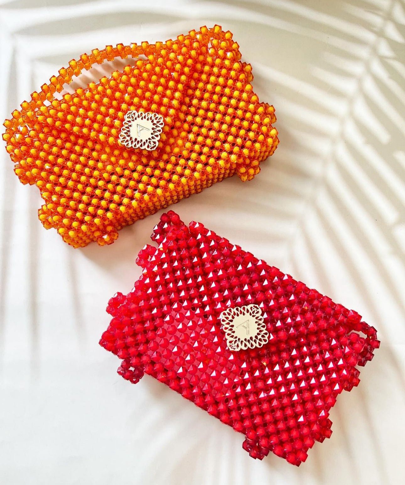 Two Ariaba Hand Beaded Clutch handbags by bibianna.co.uk—one orange, one red—feature square decorative clasps and exquisite beadwork. Handmade in Nigeria, they are displayed on a white surface with soft shadow patterns.