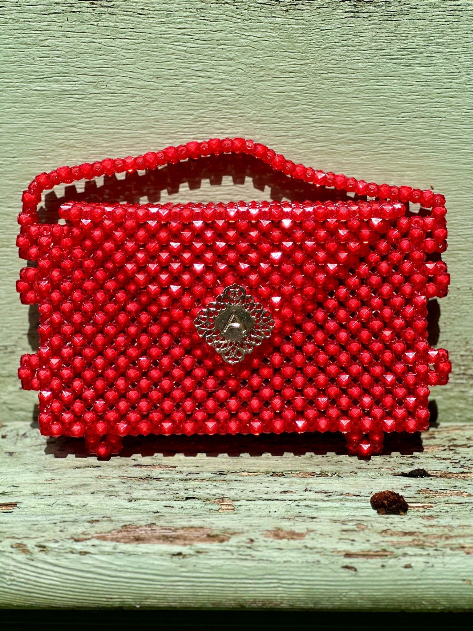 The Ariaba Hand Beaded Clutch by bibianna.co.uk is a small, rectangular bag handmade in Nigeria, featuring vibrant red beadwork and a central diamond accent. It has a handy handle and sits on a weathered pale green wooden surface.