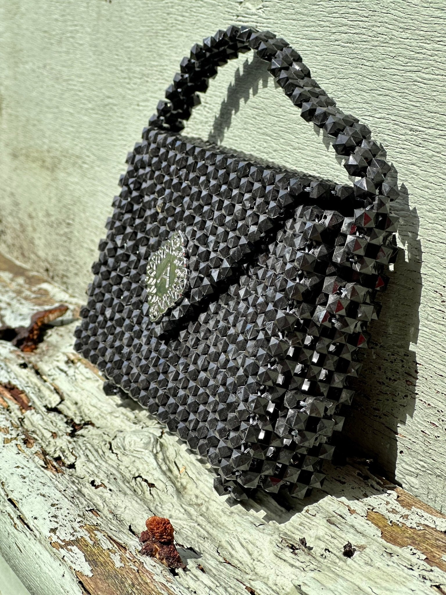 The Ariaba Hand Beaded Clutch by bibianna.co.uk features dark gray geometric beadwork, a short handle, and a decorative clasp—adding elegance to any look. It’s shown here resting on sunlit, weathered wood against a pale wall.