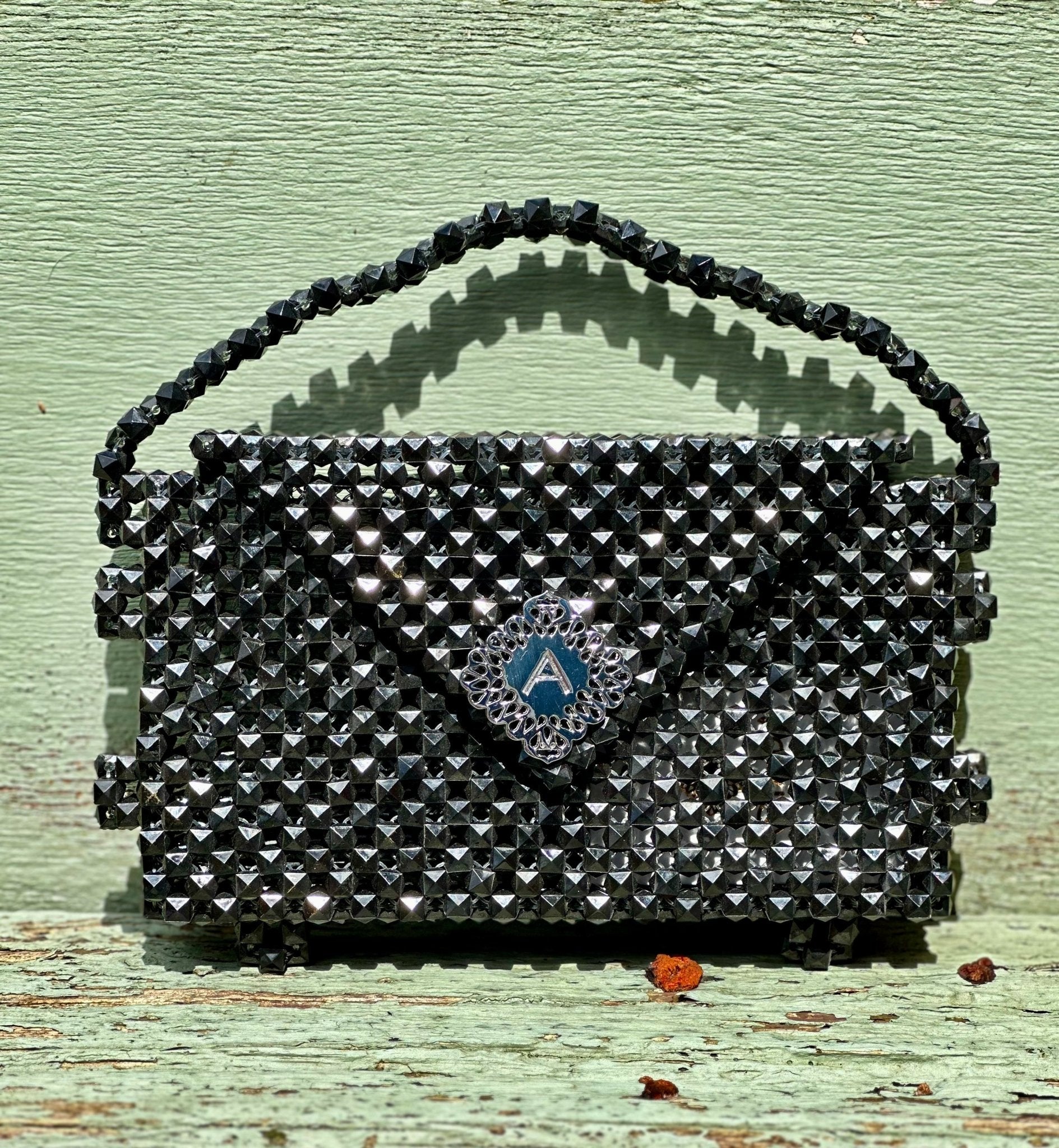 The Ariaba Hand Beaded Clutch by bibianna.co.uk is a small black handbag with intricate beadwork, a textured handle, and a blue diamond-shaped clasp featuring an "A". Displayed on pale green wood. Handmade in Nigeria.