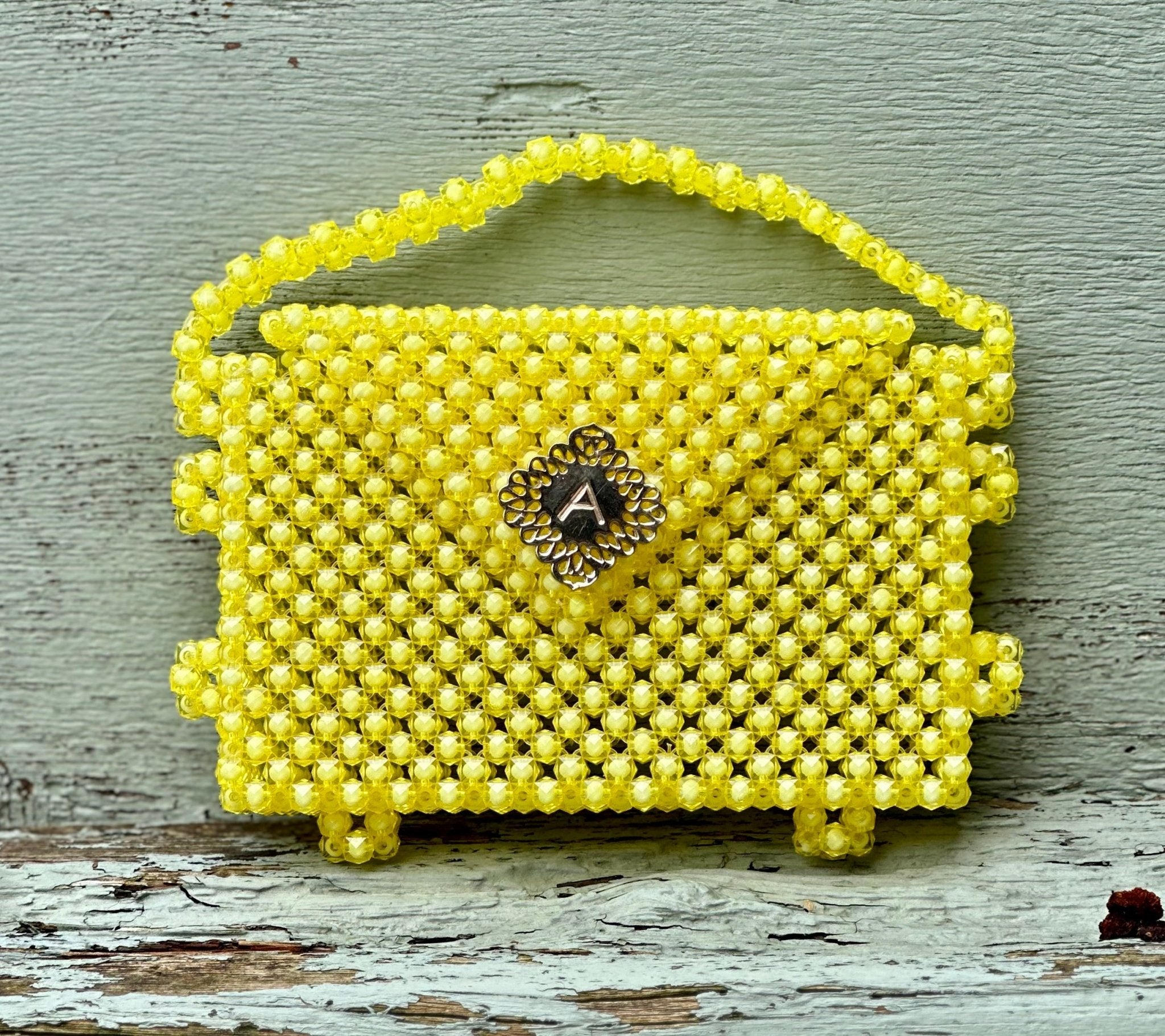 The Ariaba Hand Beaded Clutch by bibianna.co.uk is a small yellow clutch with a handle and metal clasp, showcasing intricate beadwork. Handmade in Nigeria, it is displayed against a weathered wooden background.