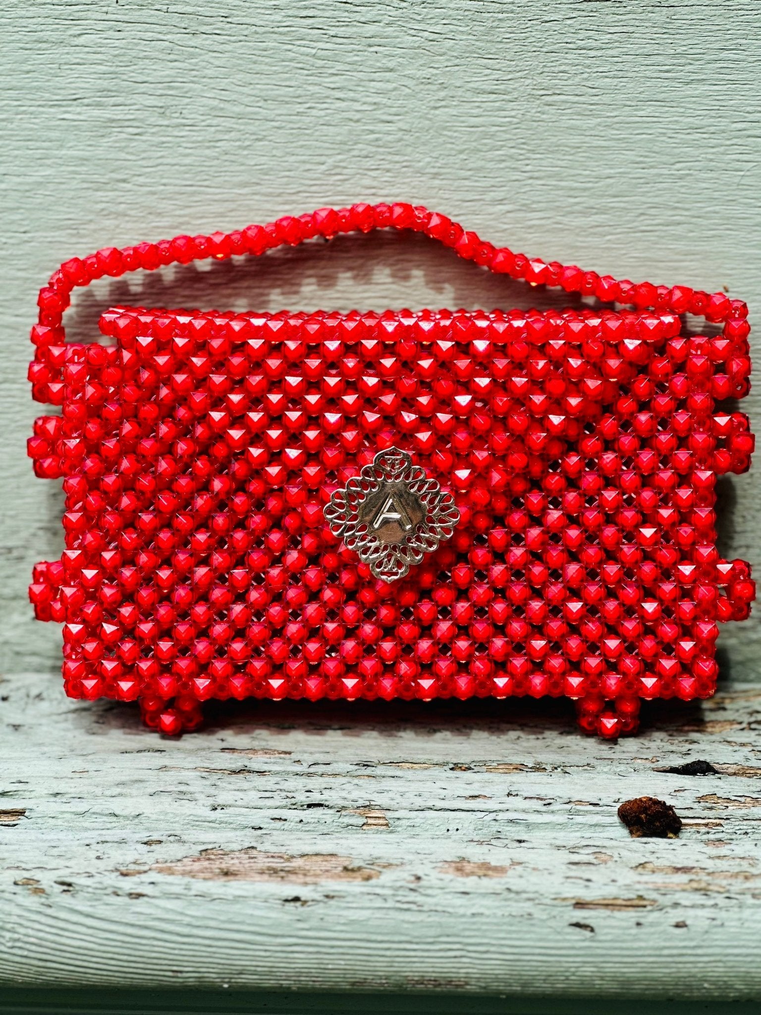 The Ariaba Hand Beaded Clutch by bibianna.co.uk features vibrant red beads and a decorative silver clasp, displayed on a pale wooden surface. Handmade in Nigeria for a unique touch.