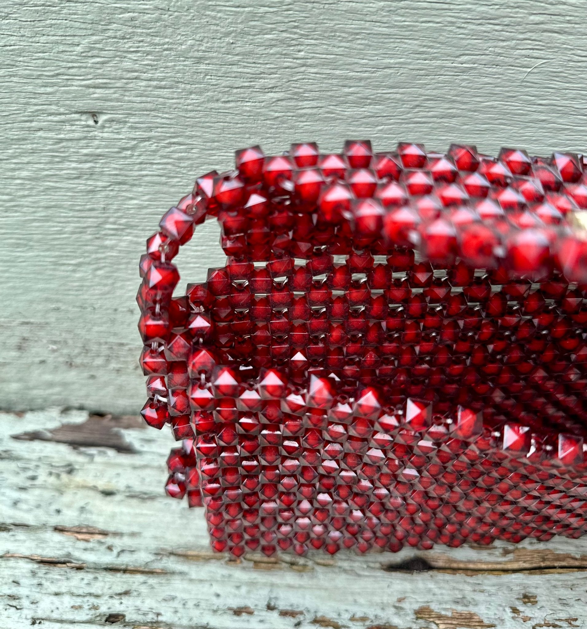The Ariaba Hand Beaded Clutch by bibianna.co.uk is a small red clutch featuring intricate hand beadwork and a handle, shown close-up on a weathered wooden surface against a light gray textured wall.