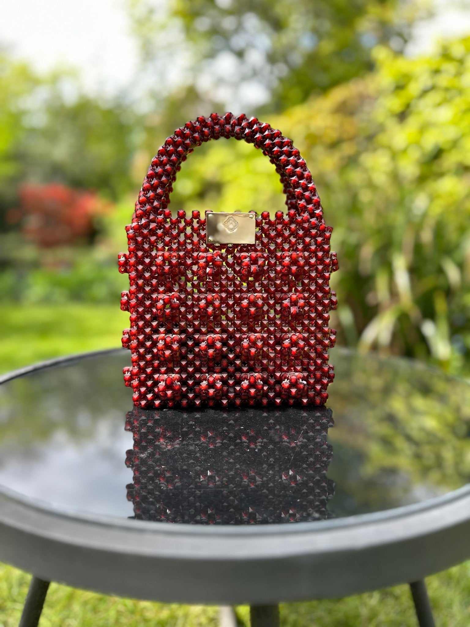 The Ariaba Ira Box Bag in Burgundy by bibianna.co.uk, featuring a handcrafted textured beaded design, rests on a glass table outdoors amid lush greenery and softly blurred plants in the background.