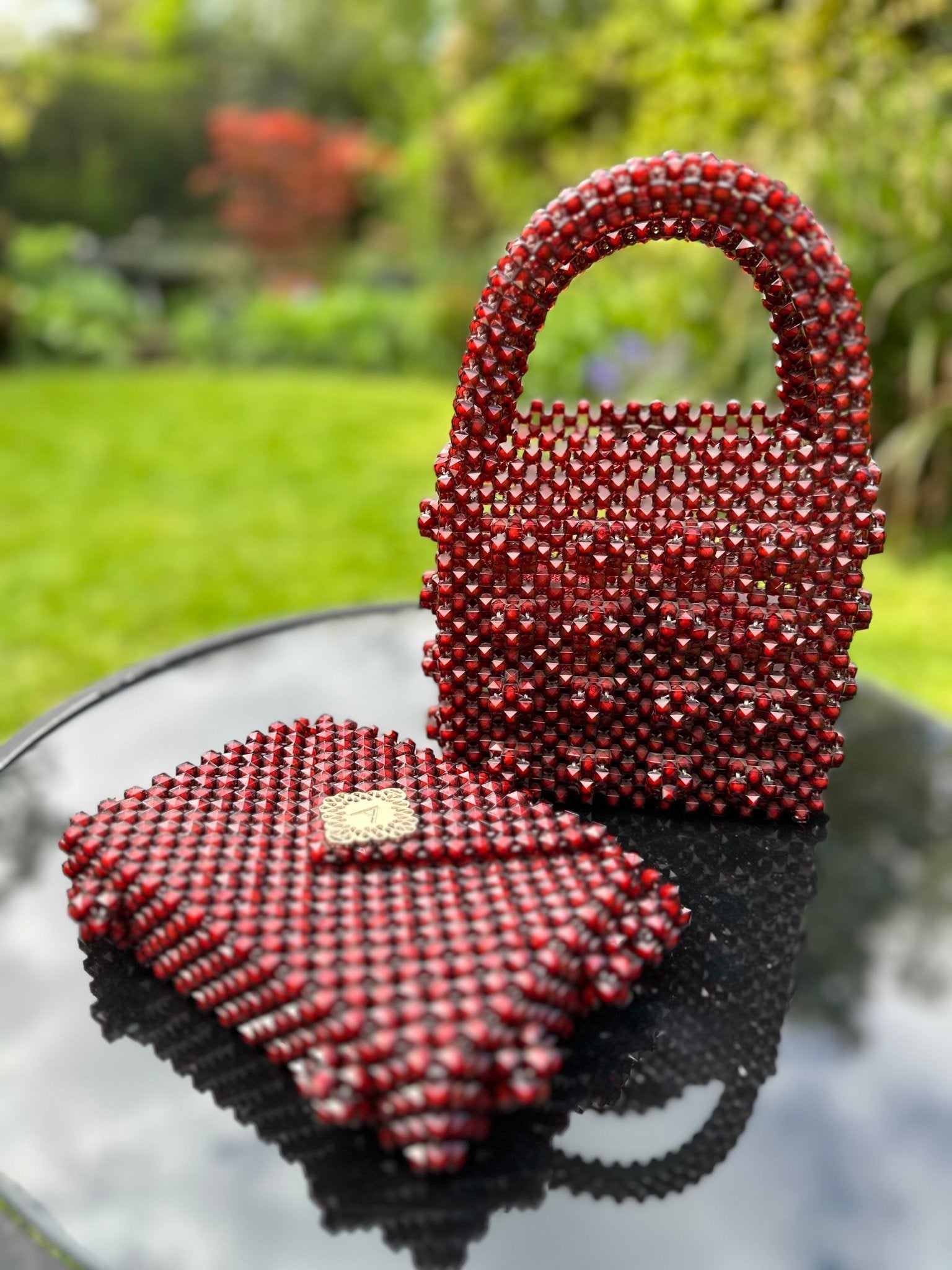 The Ariaba Ira Box Bag in Burgundy by bibianna.co.uk and a beaded clutch are showcased outdoors on a reflective black surface, with green grass and a blurred garden visible in the background.
