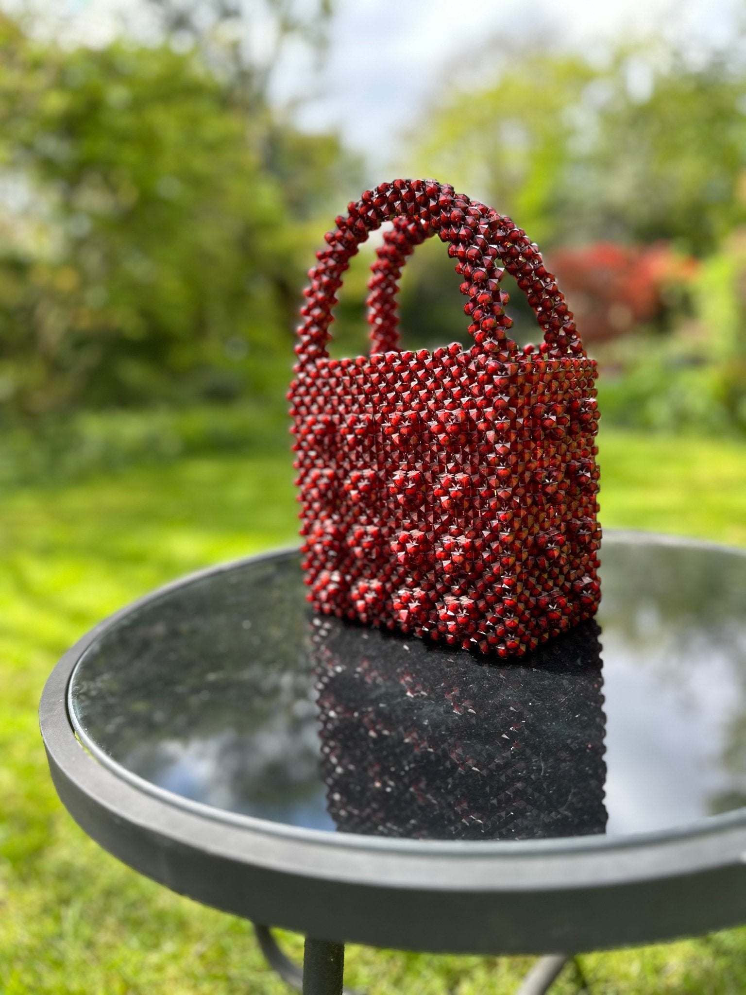 The Ariaba Ira Box Bag in Burgundy from bibianna.co.uk, a small rectangular beaded handbag with a matching handle, rests on a round glass outdoor table, its structured silhouette standing out against the green garden backdrop.