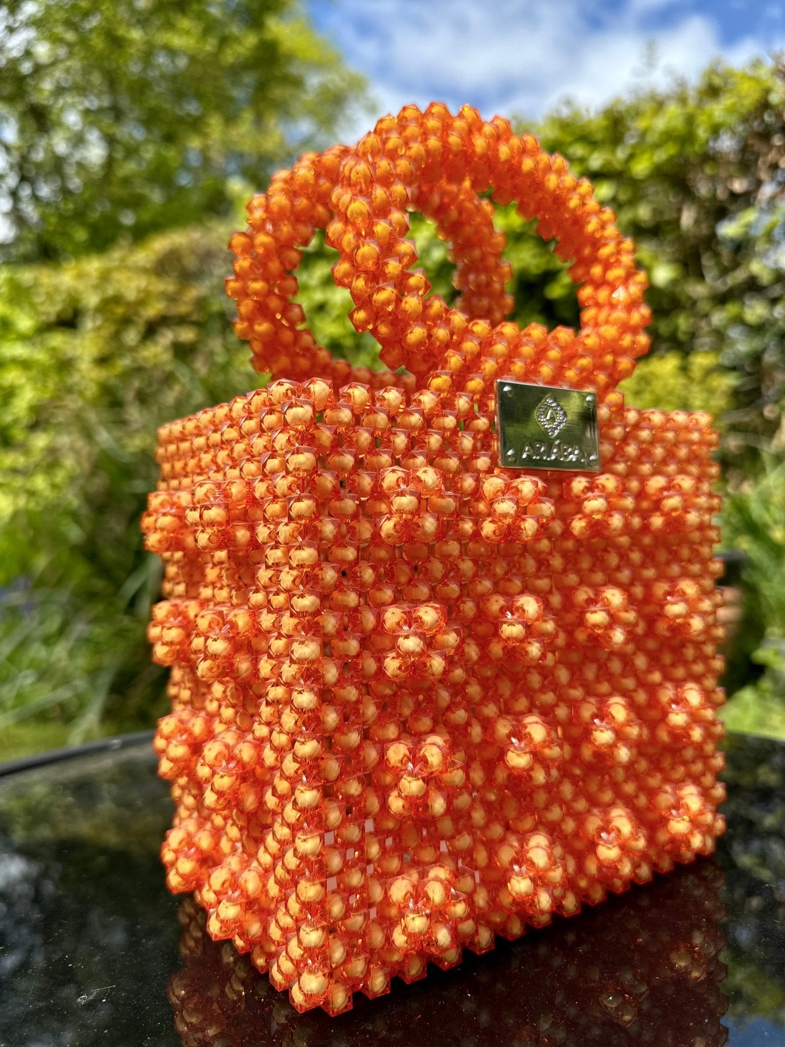 The Ariaba Ira Box Bag Orange from bibianna.co.uk, a small beaded handbag with two handles and a metallic logo, is displayed outdoors against lush green foliage and a blue sky, highlighting its handmade designer charm.