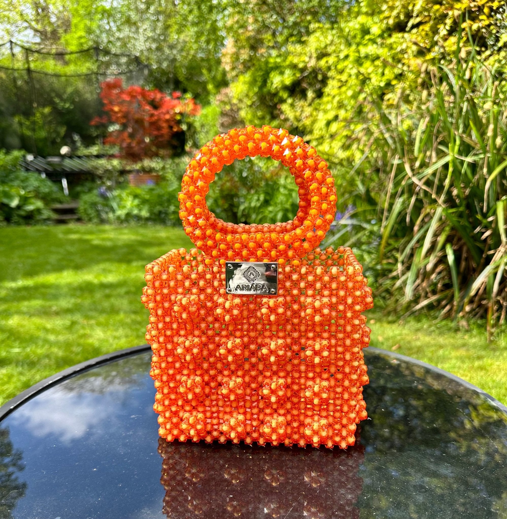 The Ariaba Ira Box Bag Orange by bibianna.co.uk, a small square beaded handbag with a silver front label, is displayed on a glass outdoor table in a lush garden—its vivid color shining in the sunlight, ideal for handmade designer bag fans.
