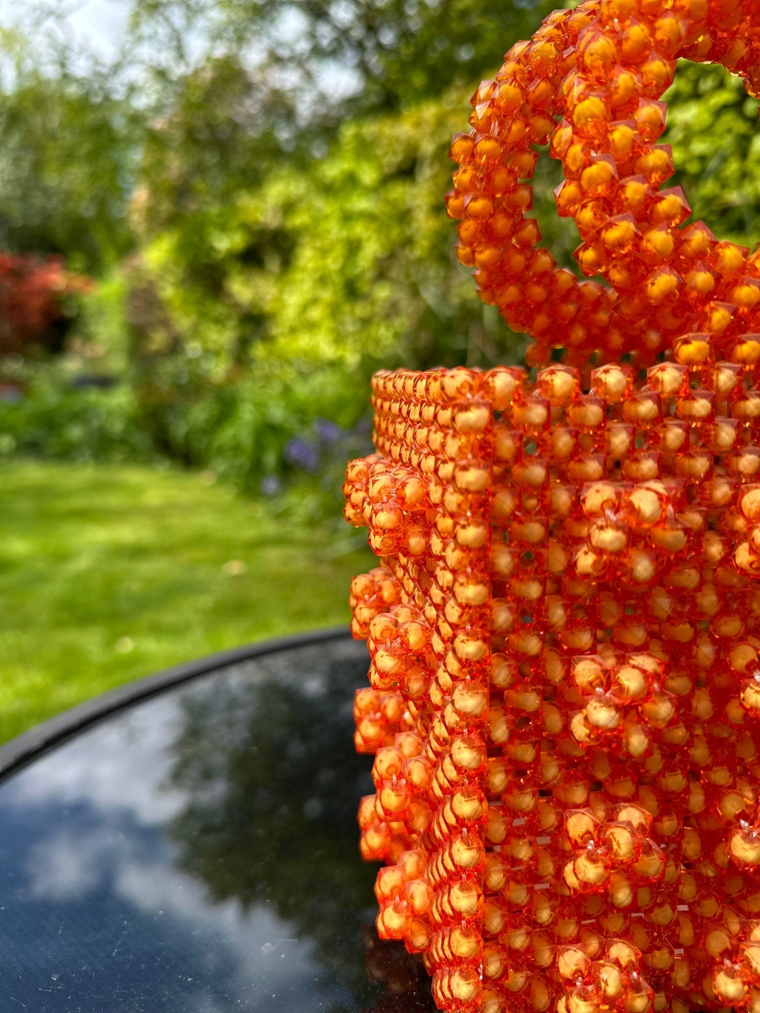The Ariaba Ira Box Bag Orange by bibianna.co.uk features striking orange beads and textured detail, showcased on a round black surface outdoors with a blurred green garden background to highlight its handmade designer appeal.