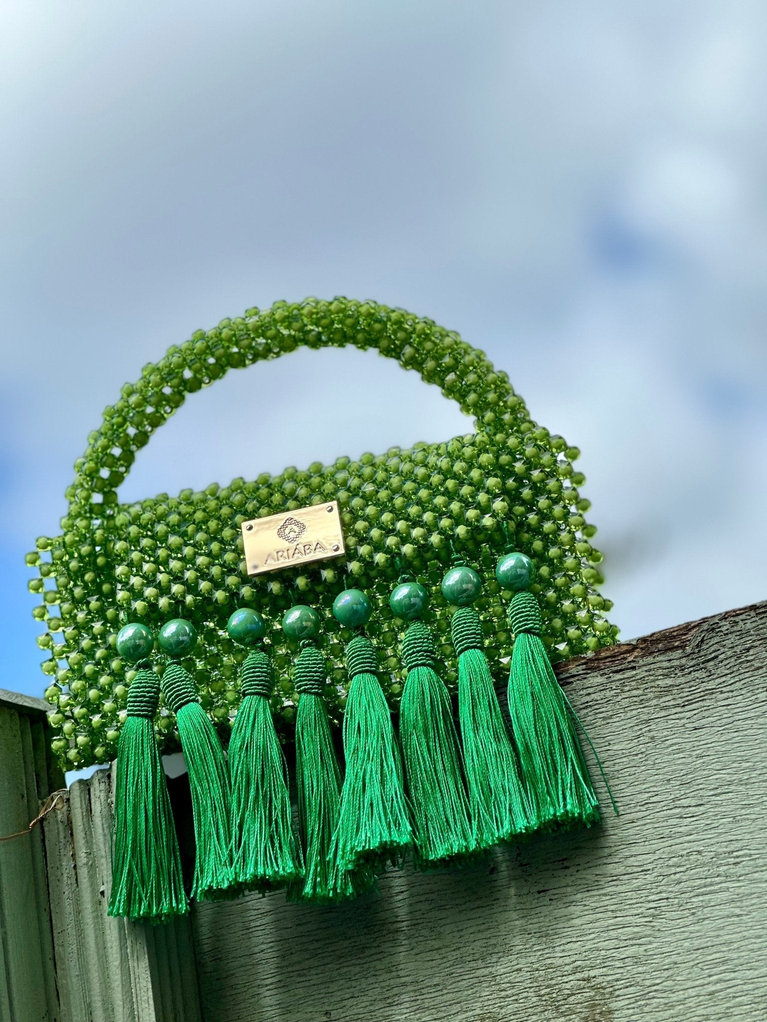 The Ariaba Ira Tassal Bag, Green by bibianna.co.uk is a handcrafted bag with green beads, short handle, gold logo plate, and green tassels—a unique Nigerian accessory.