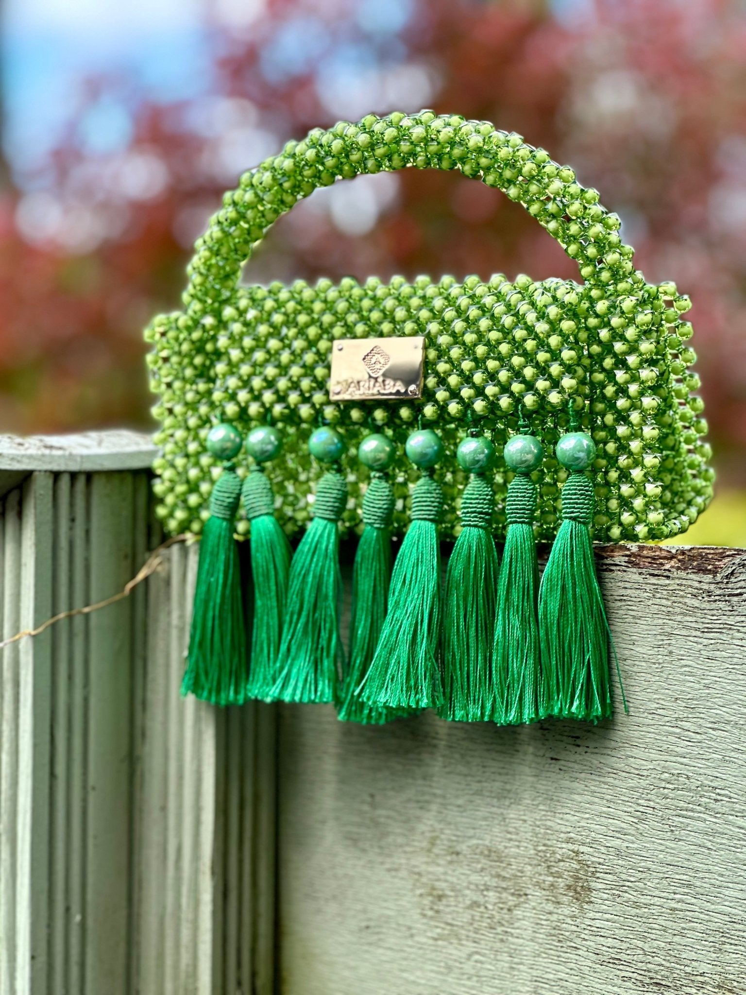 The Ariaba Ira Tassal Bag in green by bibianna.co.uk is a small handcrafted bag with a short handle, gold logo plate, and six large green tassels on the front, displayed outdoors on a wooden fence against a blurred natural background.
