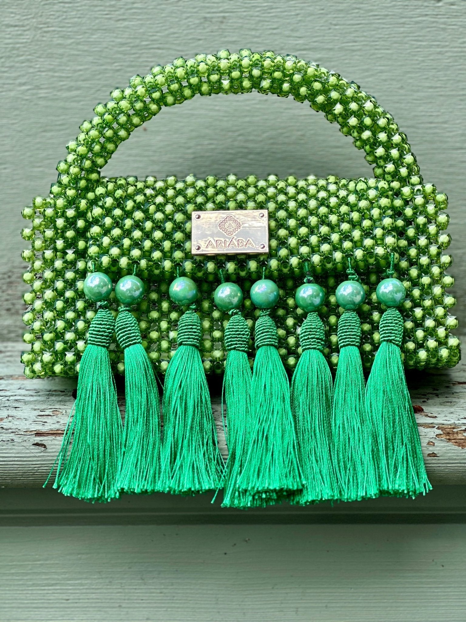 The Ariaba Ira Tassal Bag, Green by bibianna.co.uk is a handcrafted beaded handbag with a beaded handle, green tassels on the front, and a metallic label at the center—an eye-catching Nigerian accessory.