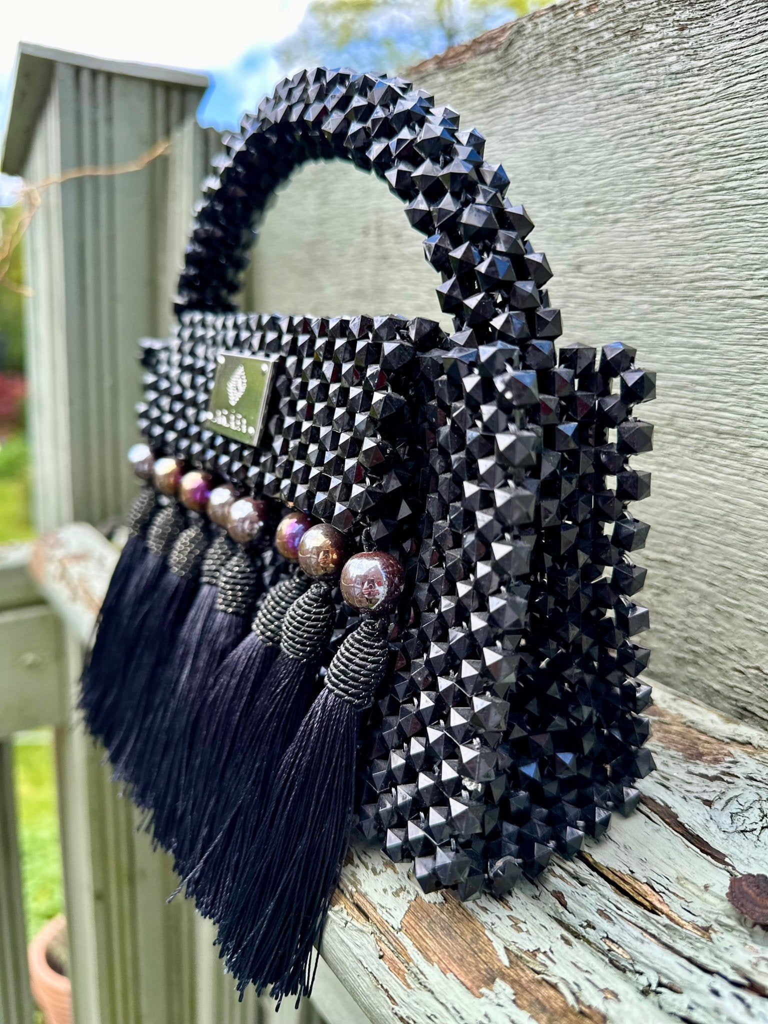 The Ariaba Ira Tasseled Bag - Black by bibianna.co.uk is a handcrafted Nigerian handbag with black beads, pearl accents, a textured studded surface, and long tassels, shown on a weathered wooden railing outdoors.