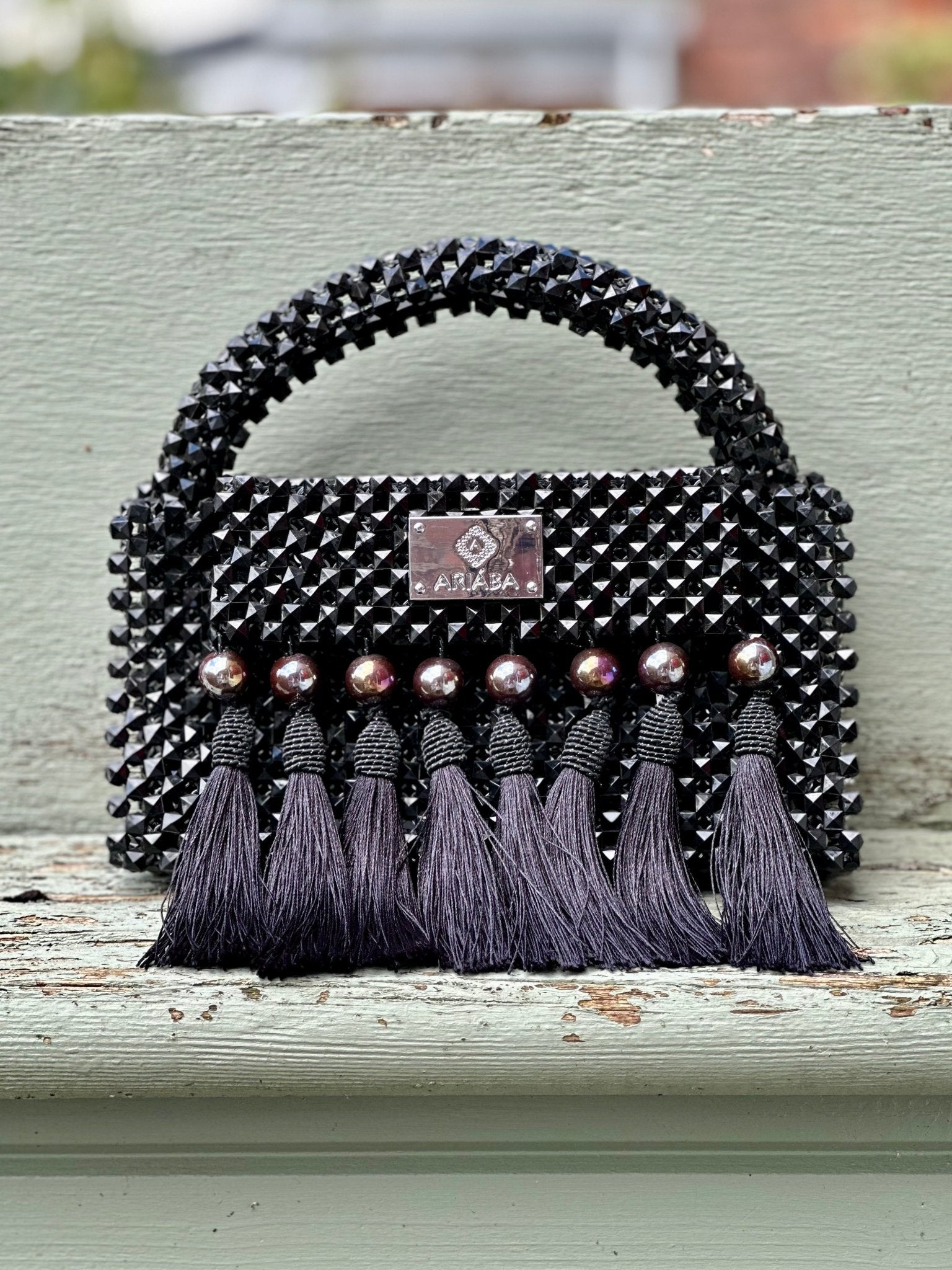 The Ariaba Ira Tasseled Bag - Black by bibianna.co.uk features a beaded handle, silver brand plaque, and six silver beads with long tassels. Handcrafted in Nigeria, it's a striking accessory shown on a light green wooden surface.