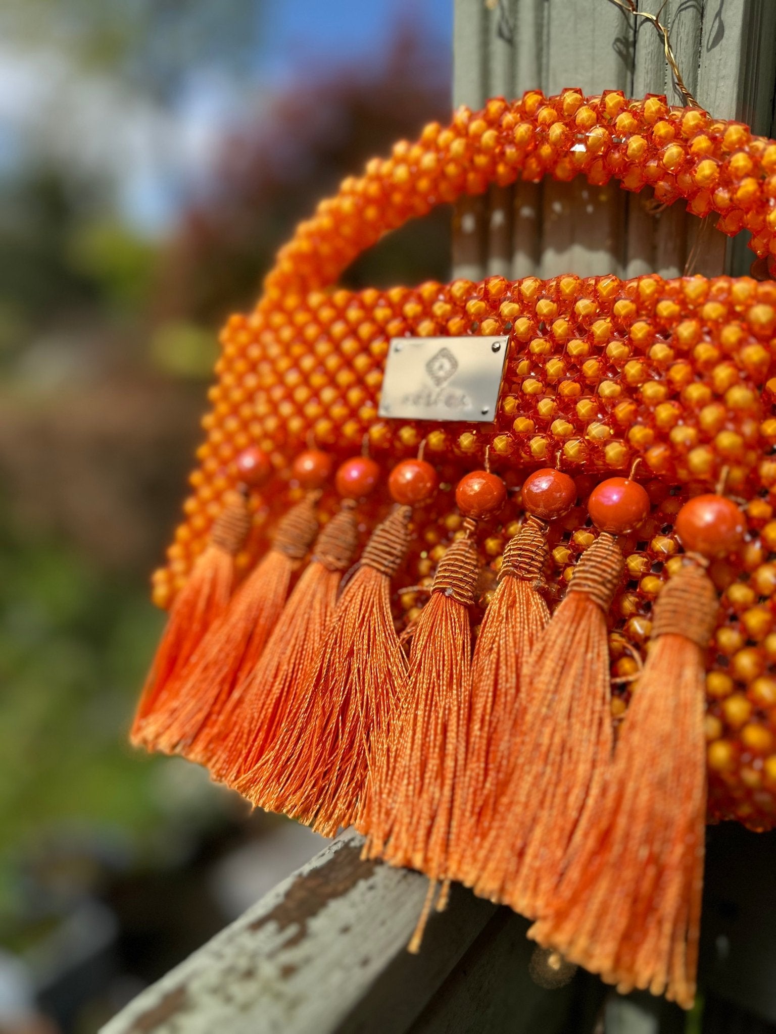 A close-up of the Ariaba Ira Tasseled Bag in orange by bibianna.co.uk, handcrafted in Nigeria with bold beads and tassels, set against a softly blurred outdoor backdrop of greenery and wood.
